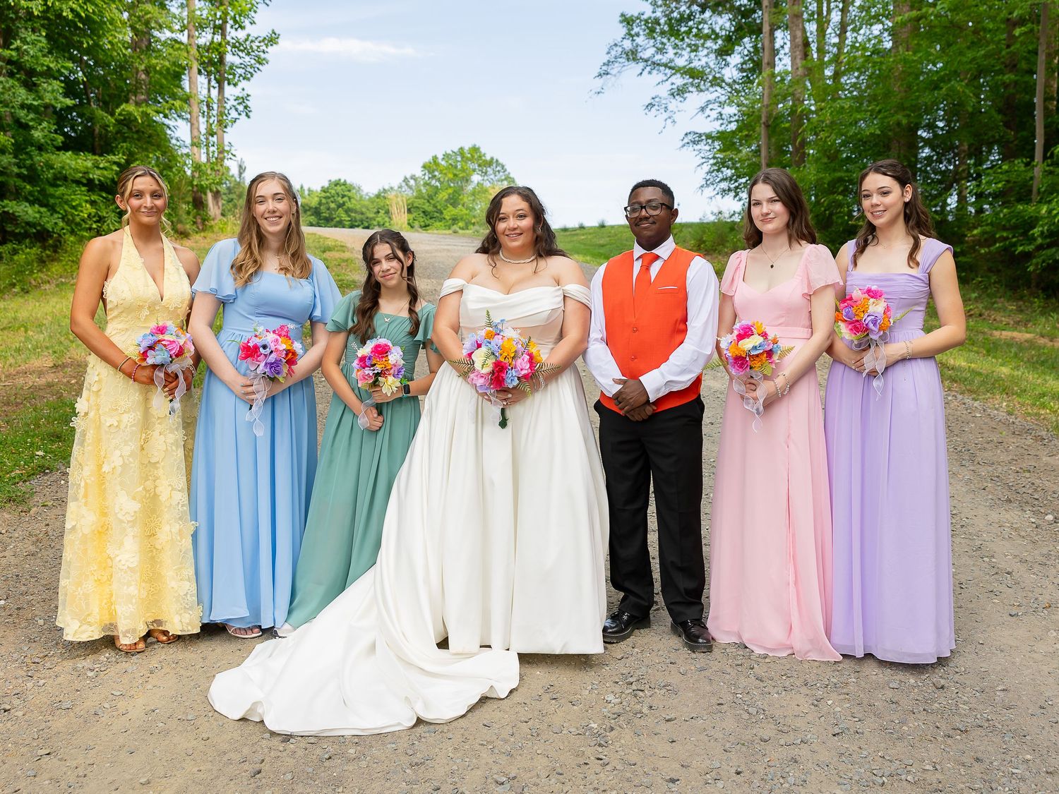 Wedding party posing outdoors, bride in white gown with groom and five bridesmaids in colorful dresses holding bouquets.
