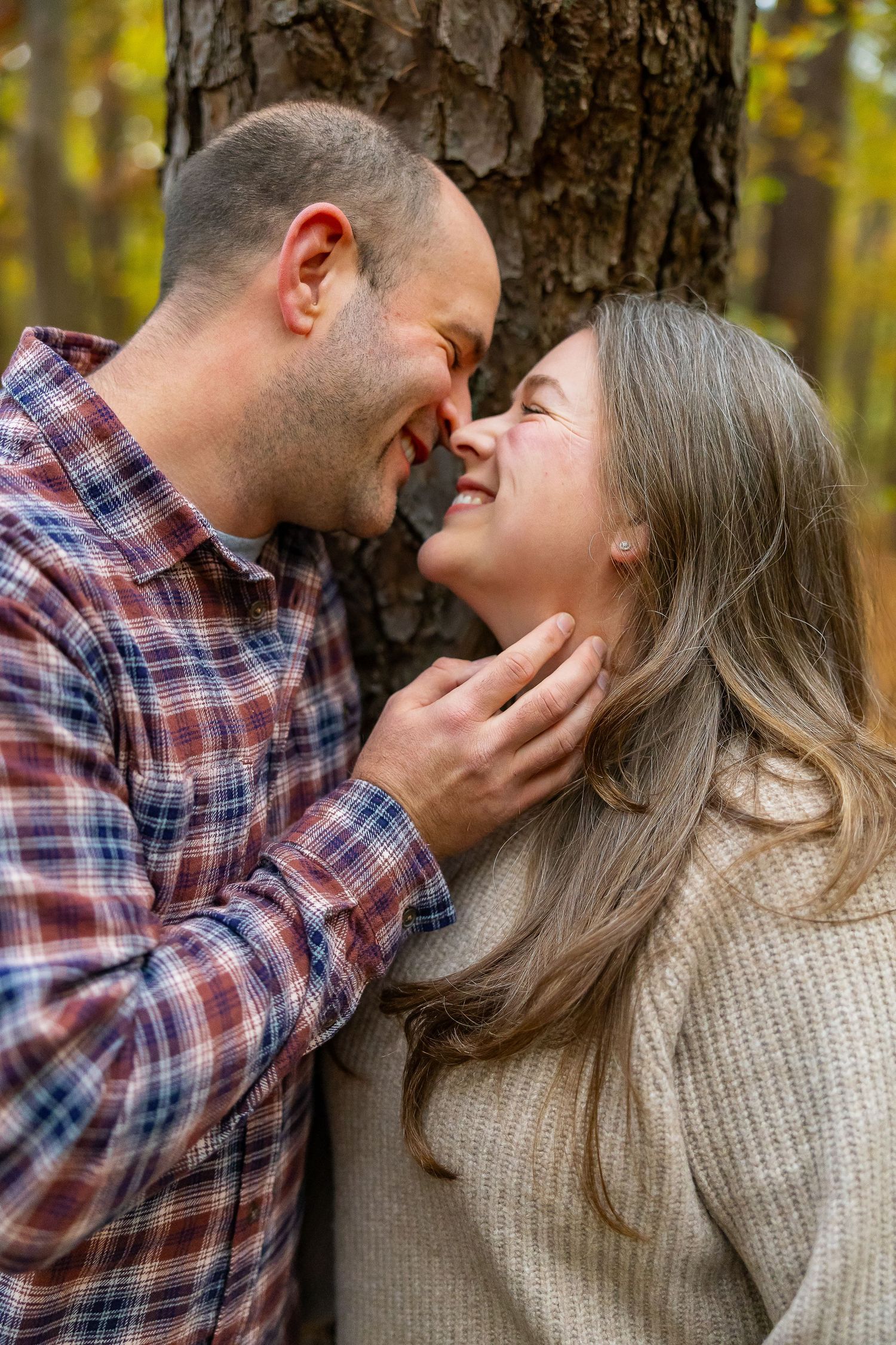 A romantic couple shares an intimate moment leaning against a tree trunk in an autumn forest setting.