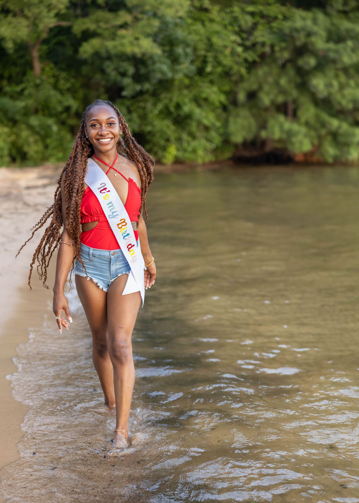 A sash-wearing woman wades through shallow river water while smiling in a red swimsuit and denim shorts.
