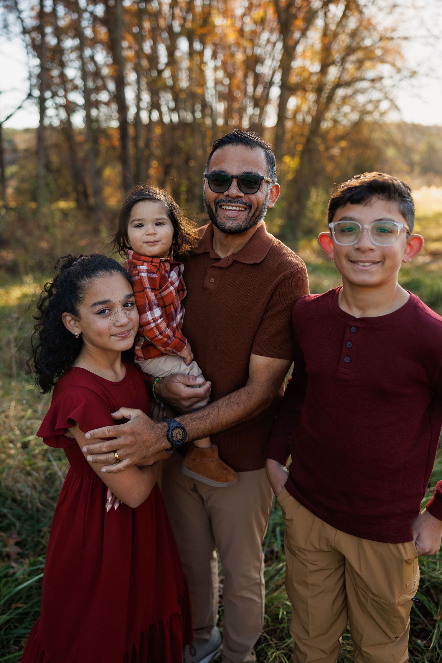 A family wearing burgundy and brown clothing poses together for outdoor autumn portraits in a wooded setting.