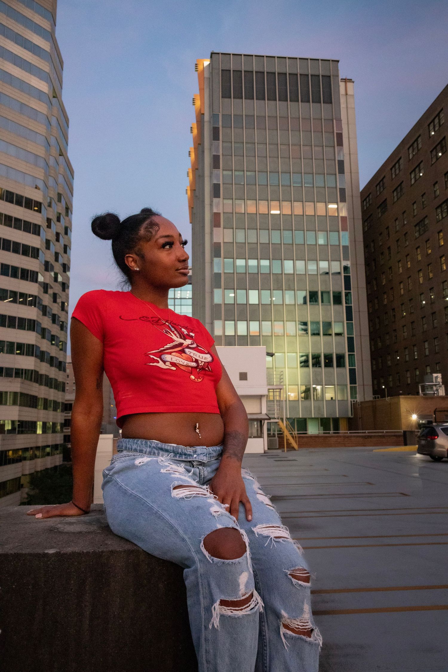 Urban fashion portrait with red crop top and distressed jeans against city buildings at sunset.