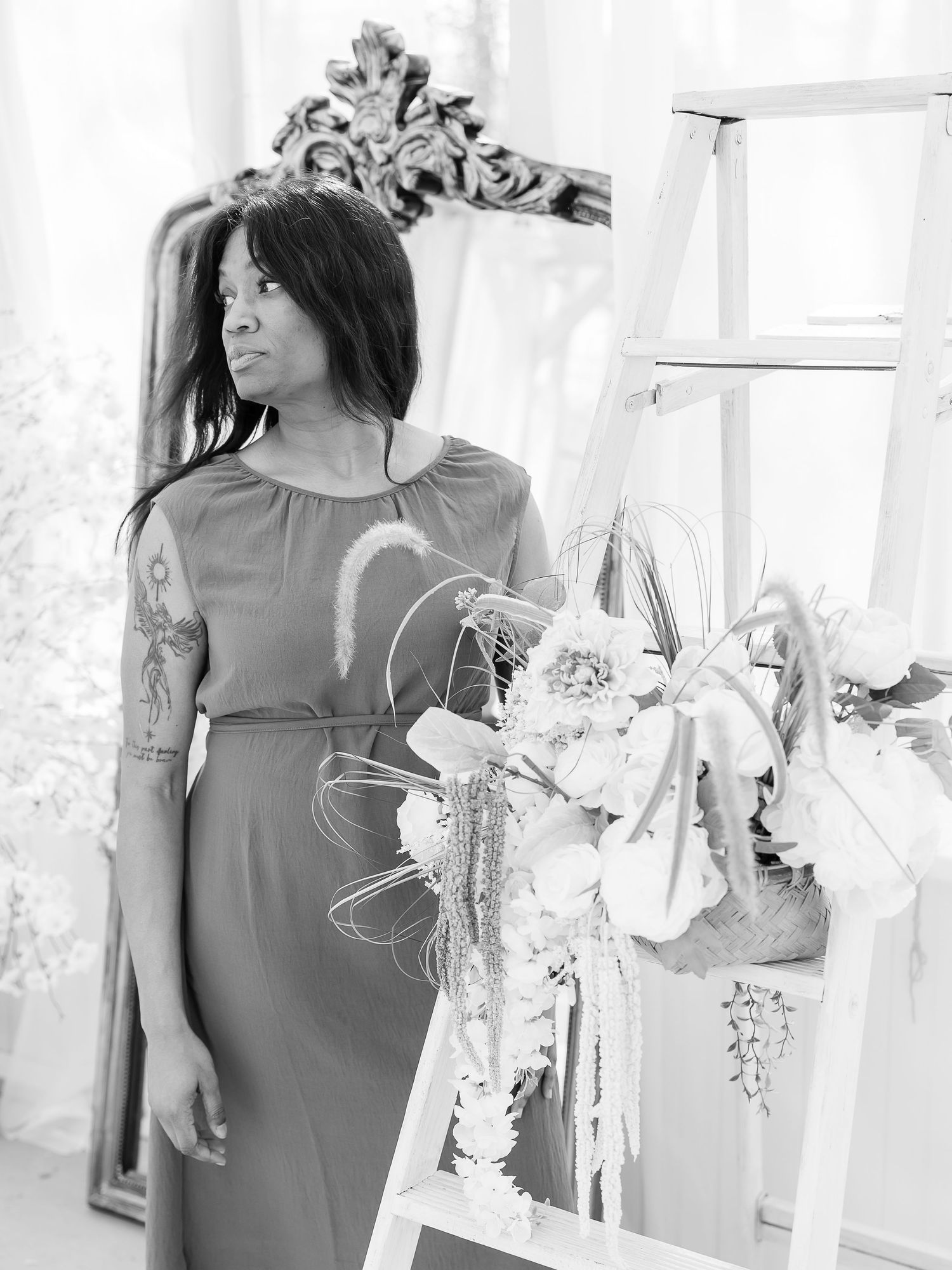 Black and white photo of a woman in a dress holding a floral bouquet near a decorative mirror and white ladder.