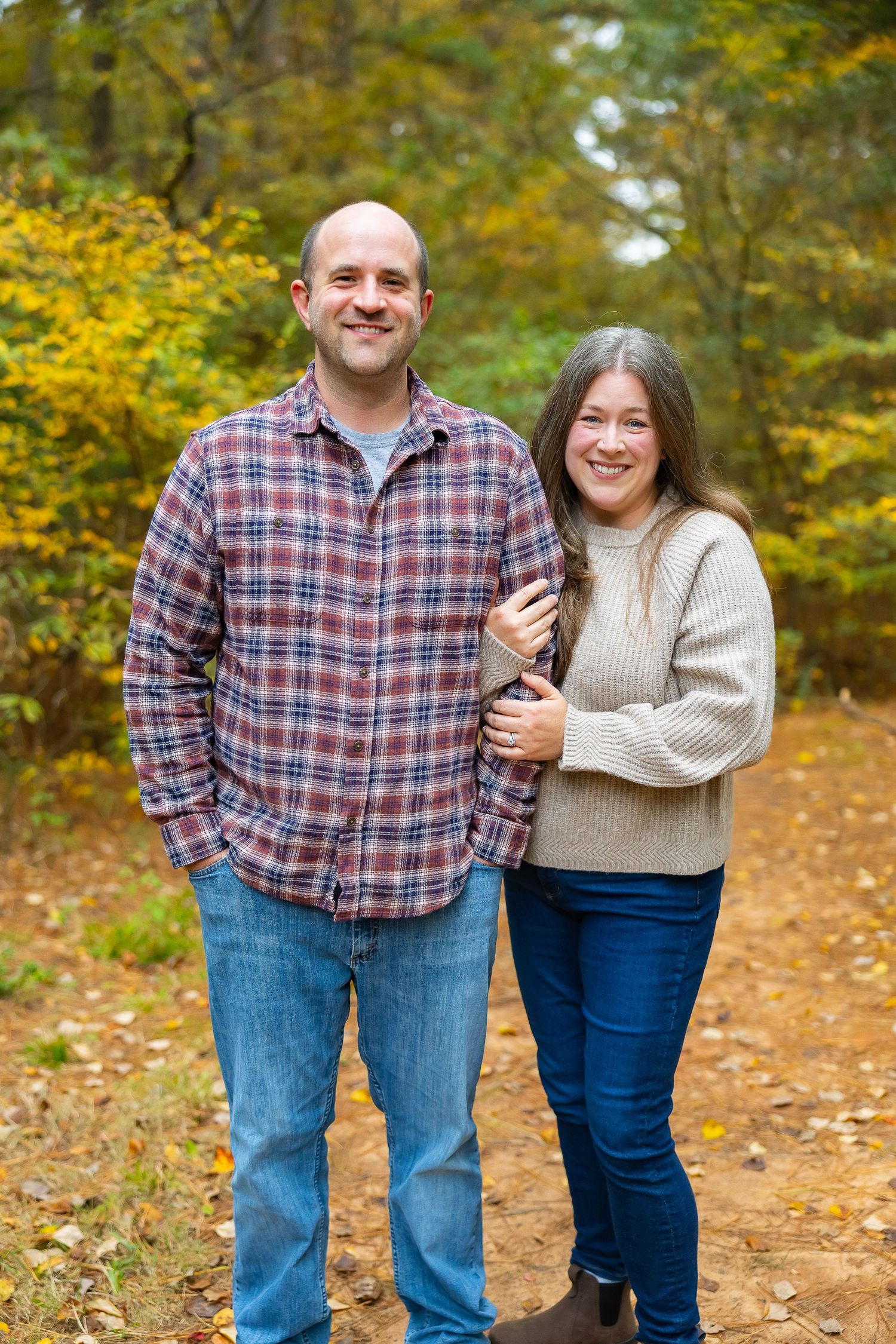 A couple stands together on a leaf-covered autumn path surrounded by yellow and green foliage in the background.