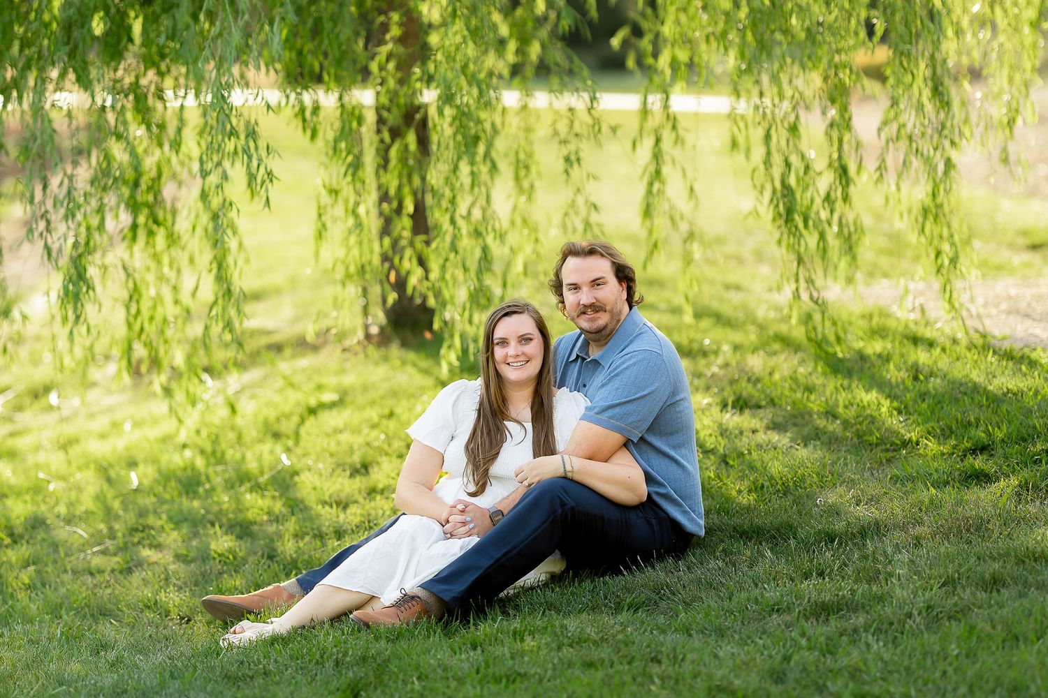 Couple sitting together on lush green grass under a weeping willow tree outdoors.