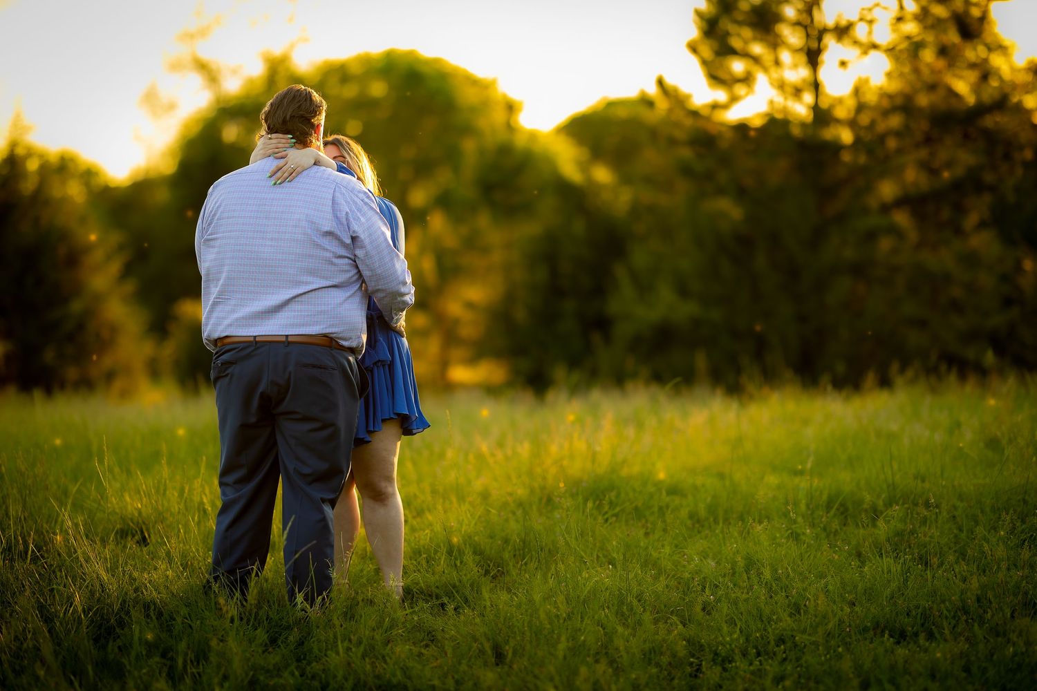 Couple embracing in a sunny green meadow at golden hour with trees in the background.