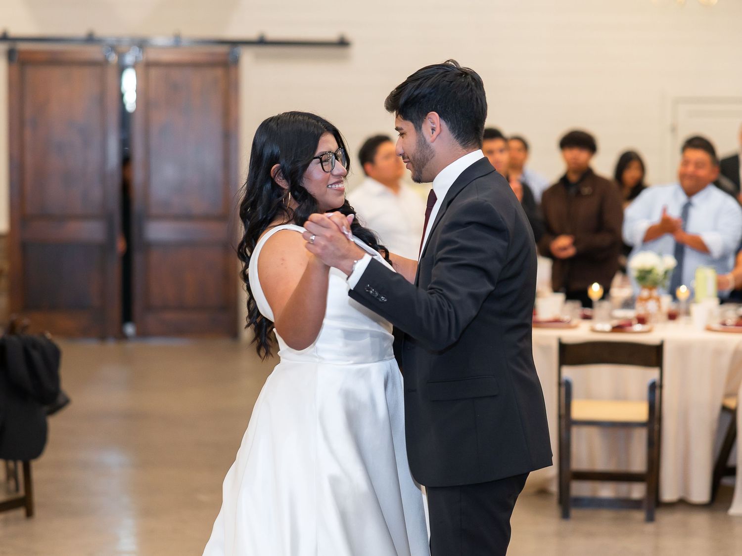 Bride and groom share their first dance at wedding reception, guests watching in elegant barn venue.
