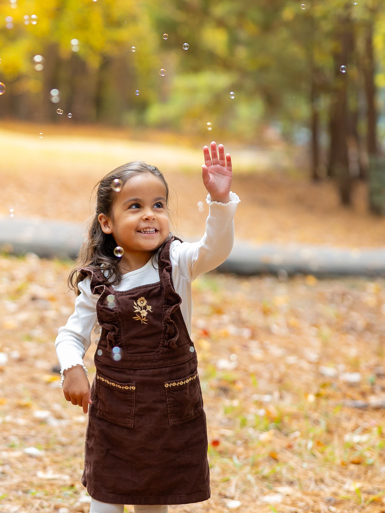 A young child in a brown dress plays joyfully among scattered autumn leaves in a sunlit outdoor setting.