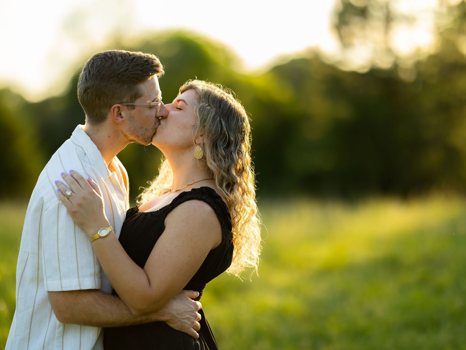 A couple shares a romantic kiss in a sunlit green field during golden hour outdoors.