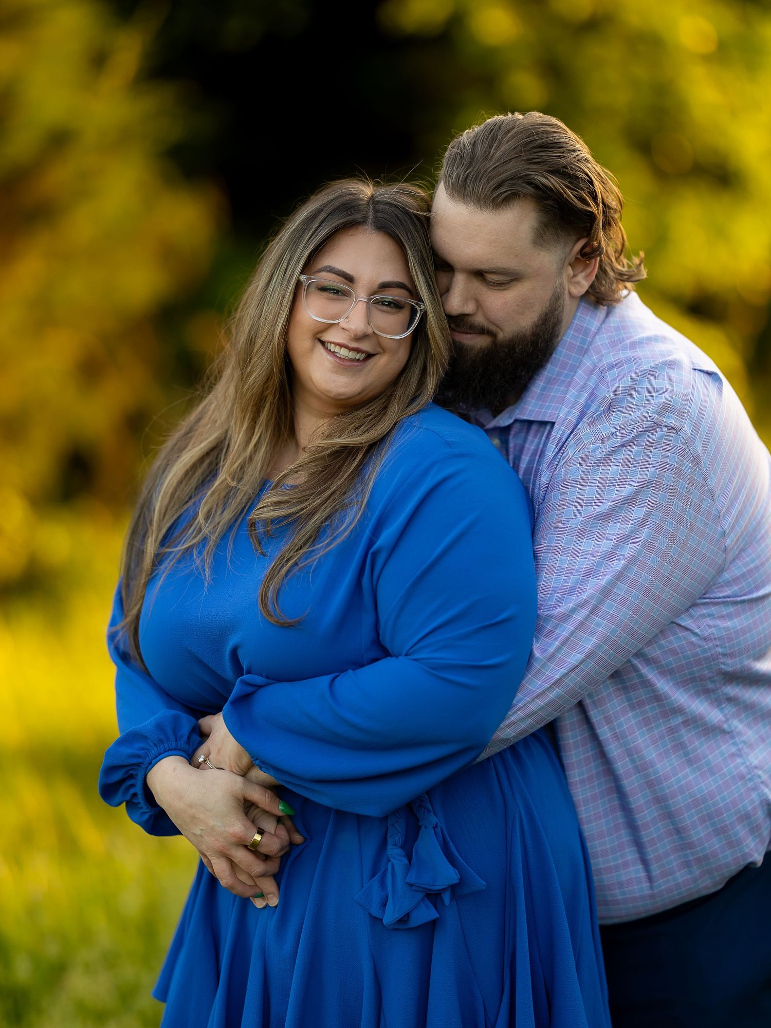 Couple embracing outdoors, woman in blue dress smiling, man with beard kissing her cheek, golden foliage background.