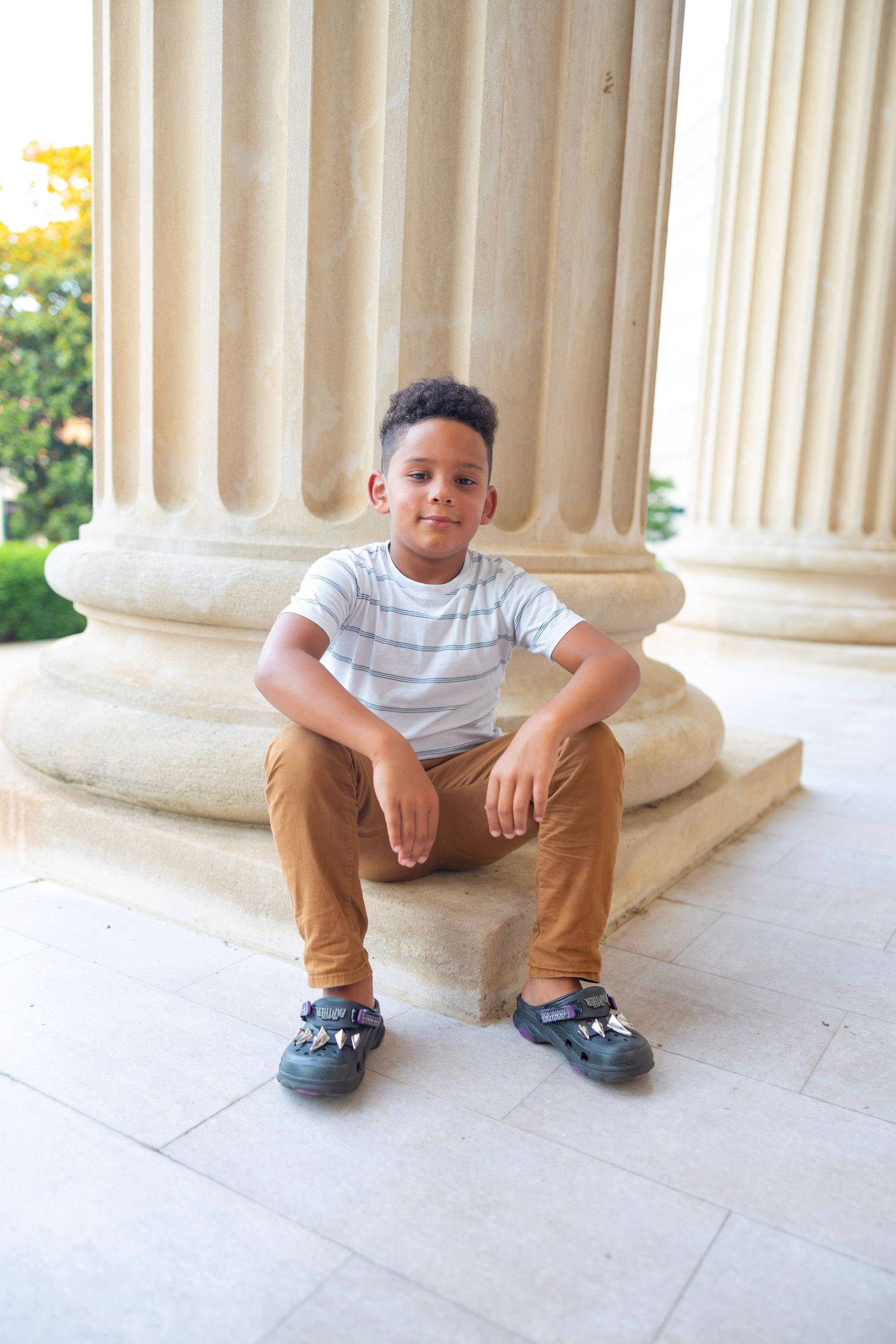 Young boy in striped shirt and tan pants crouches down between white columns on a stone porch.
