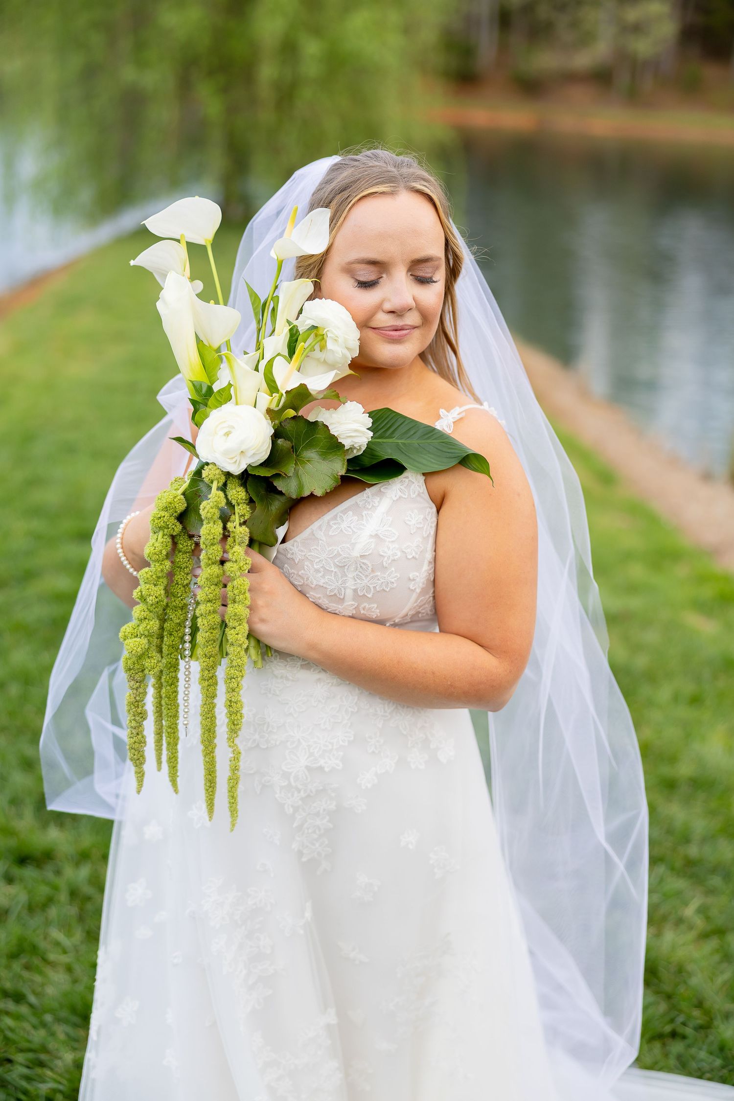Bride in white lace gown and veil holds white calla lily bouquet with green accents, eyes closed, near a pond.