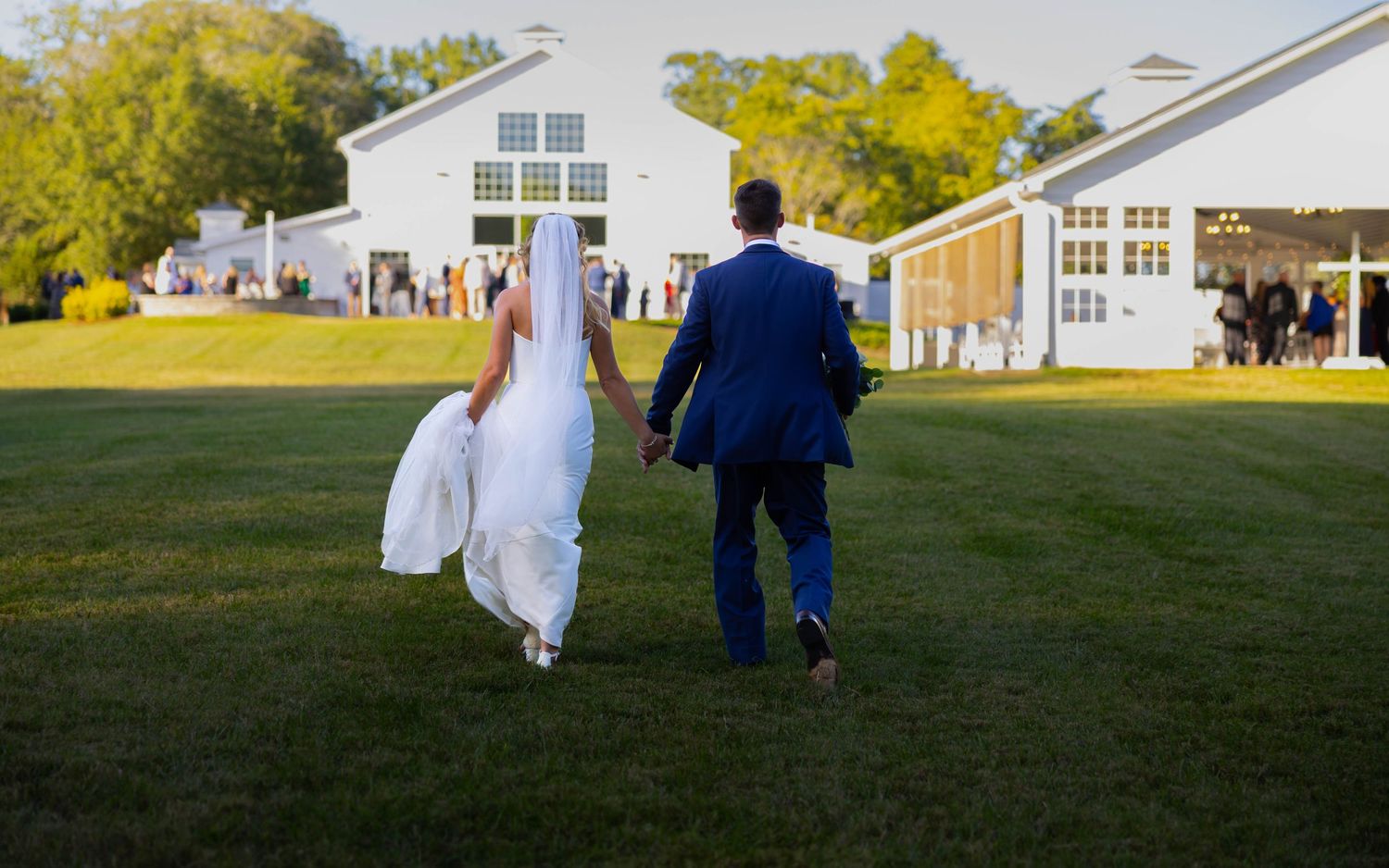 Bride and groom walking hand in hand across green lawn toward white wedding venue on sunny day.