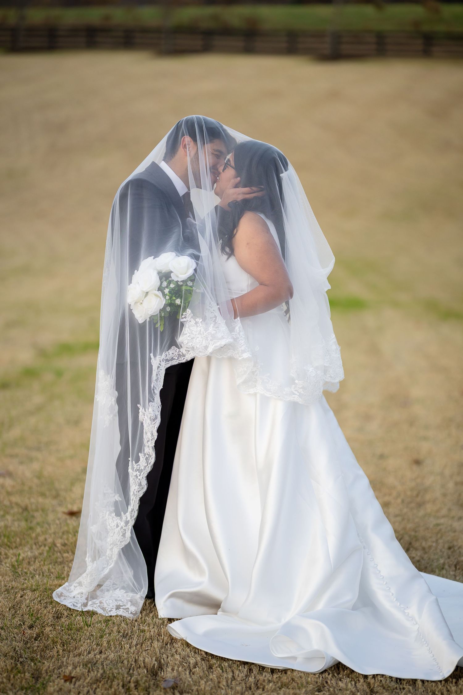 Bride and groom kissing under wedding veil outdoors, bride holding white bouquet on grassy field.