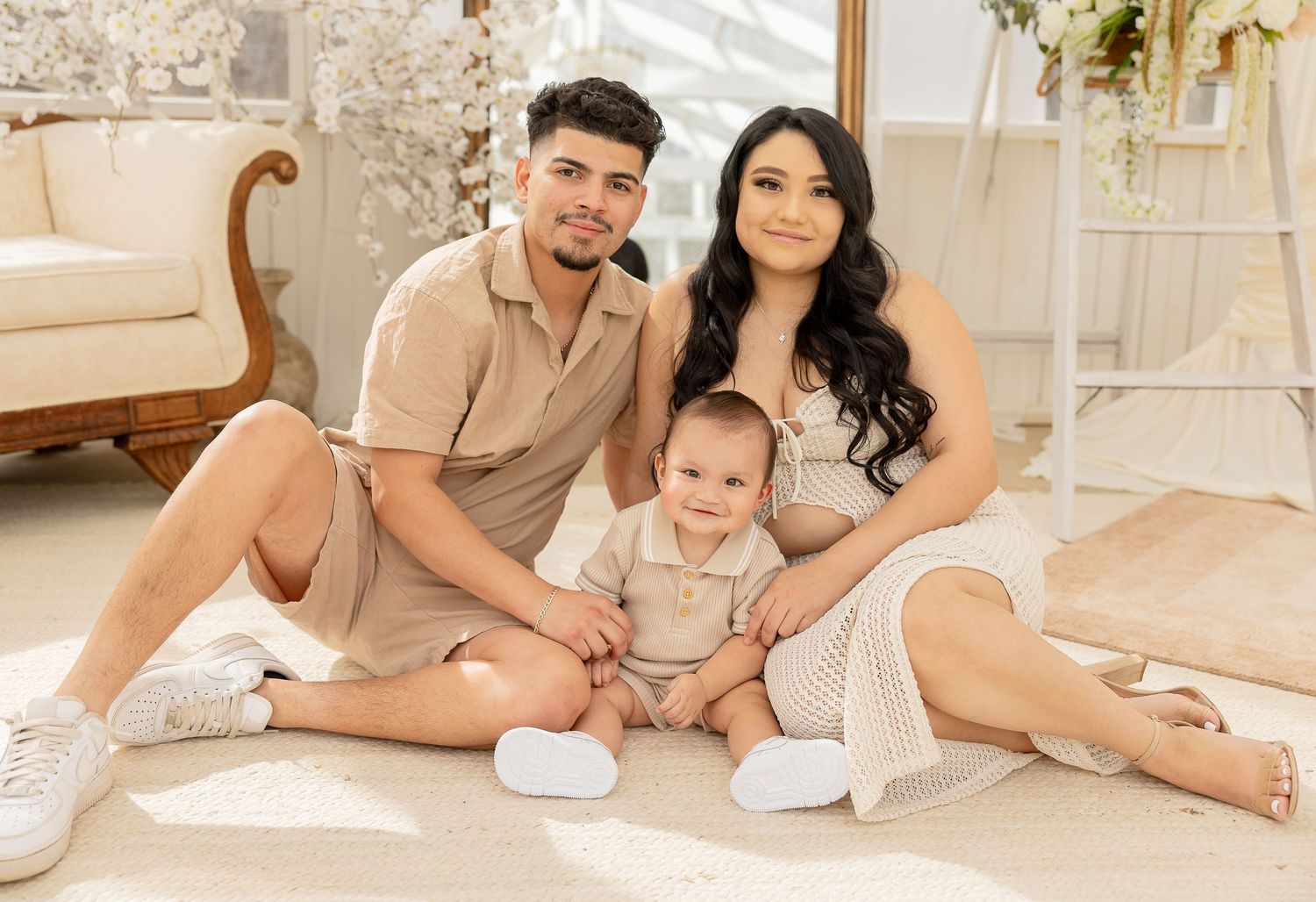 Young family portrait with parents and baby sitting on floor in bright white studio setting.