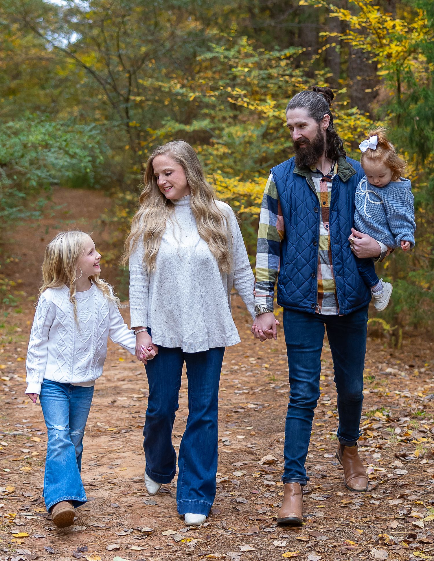 A family in matching white tops and denim walks together on a leaf-covered forest path in autumn.