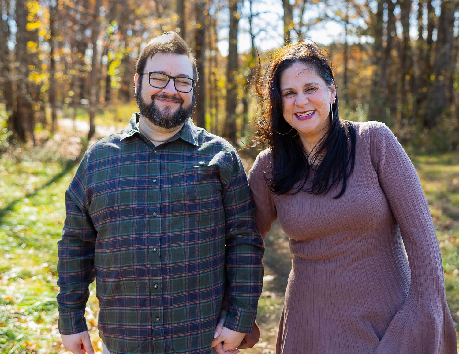 A couple in fall attire smiles together during an outdoor autumn photoshoot in a wooded setting.