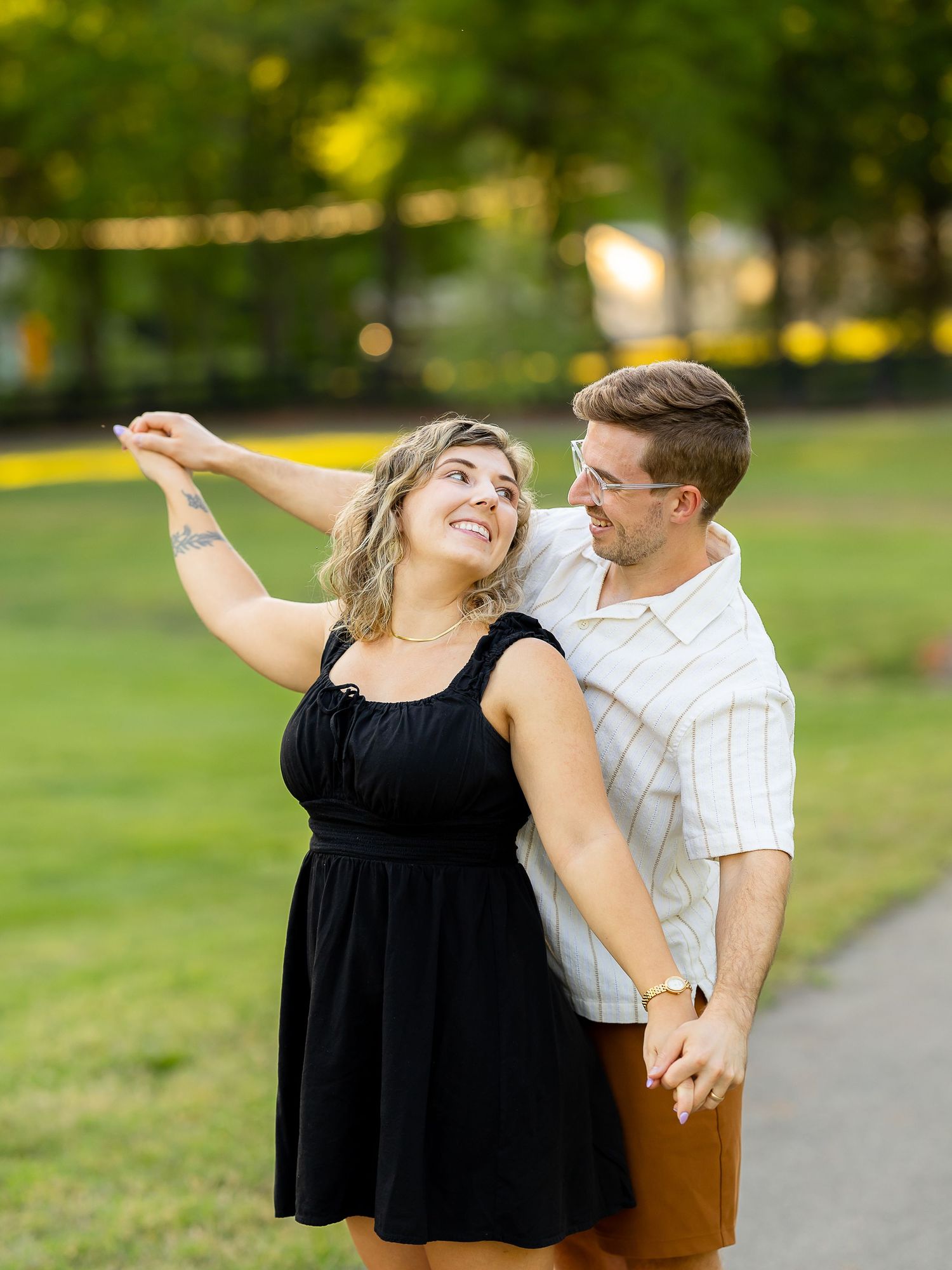 A smiling couple holds hands and dances together in a sunny park, the woman in a black dress with a tattoo on her arm.