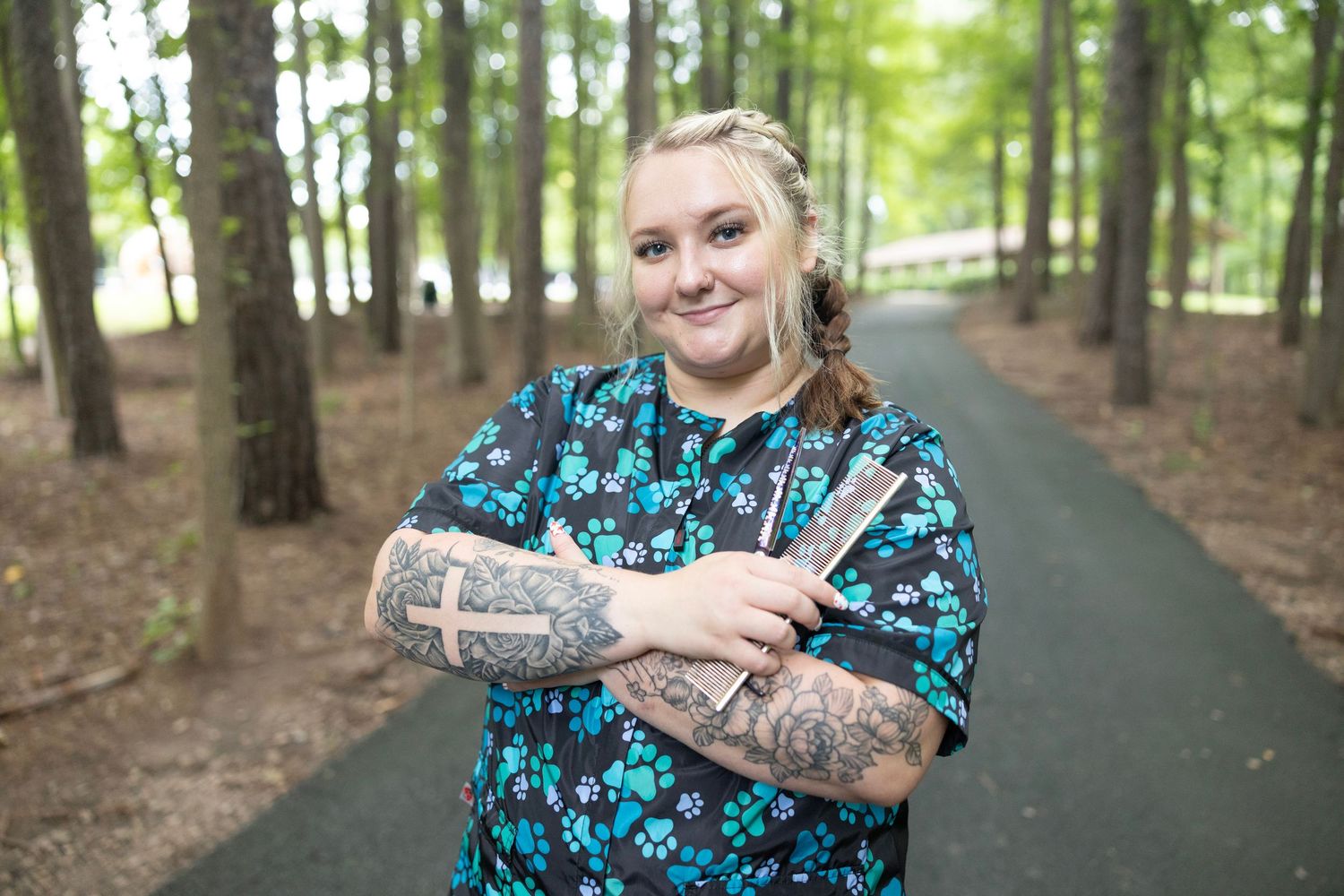 A person with tattoos wearing a blue floral print top stands on a paved path in a wooded area.