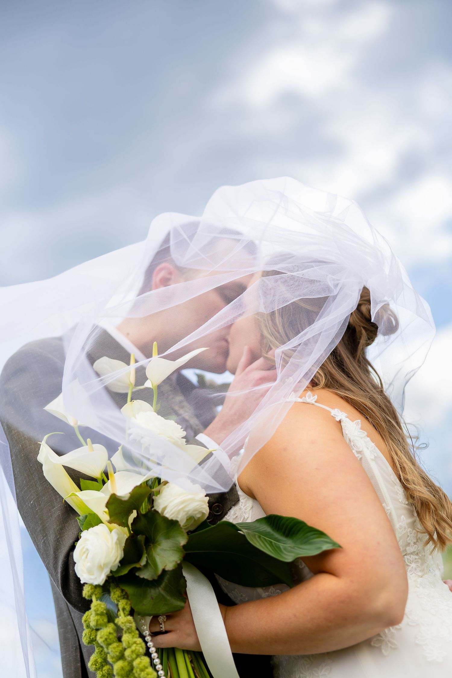 Bride and groom sharing a kiss under a flowing white veil, holding a bouquet of white calla lilies outdoors.