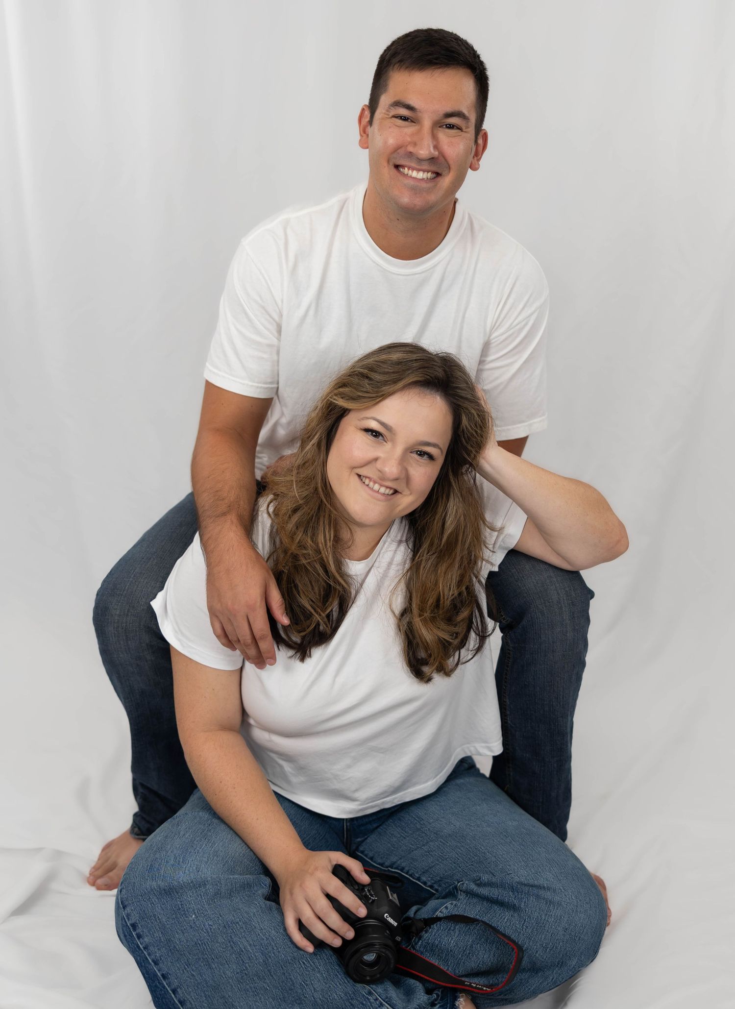Happy couple in casual white shirts and jeans sitting and smiling together during a playful studio photoshoot.