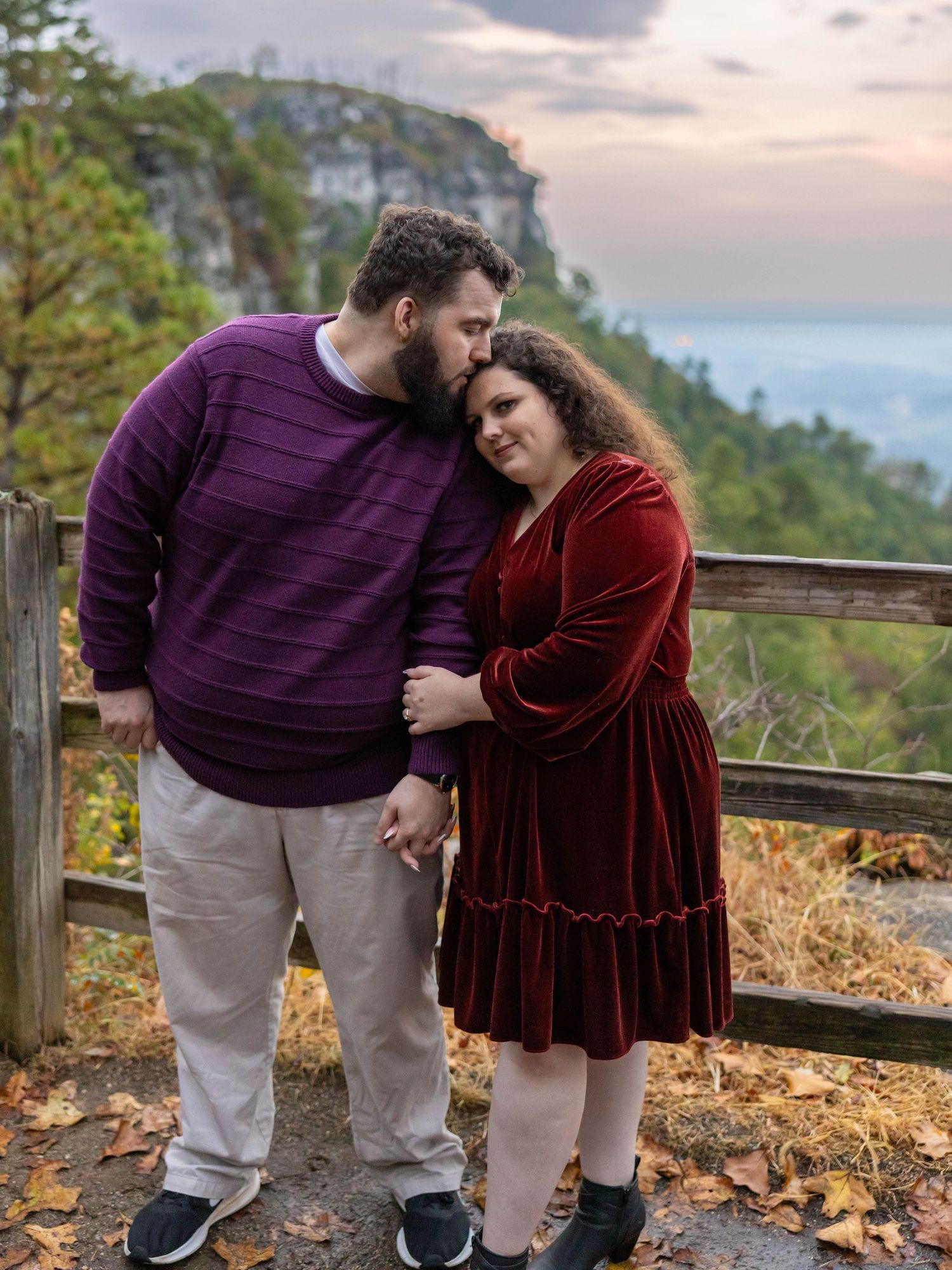 A couple in casual attire shares a tender moment while standing on an autumn hiking trail during sunset.