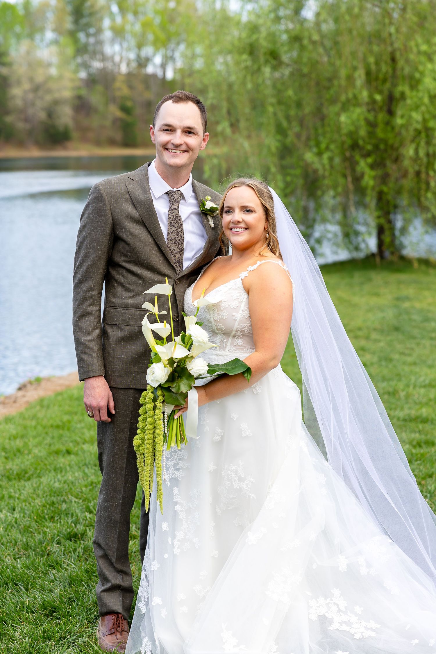 Bride and groom posing outdoors by a lake, bride holding white floral bouquet with veil, groom in brown suit.