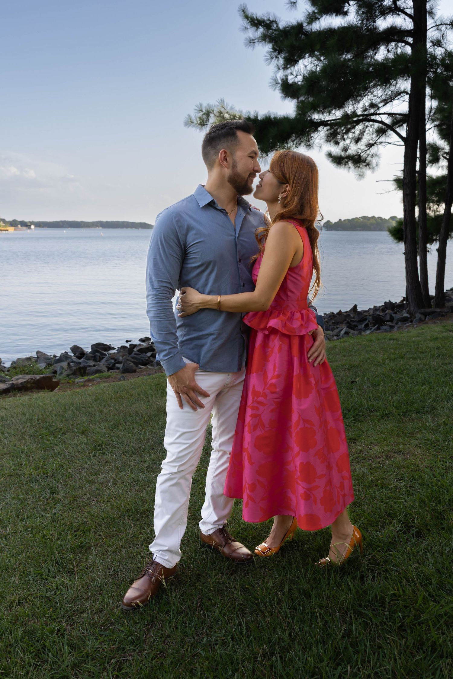 A couple embraces by a lakeside at dusk, with her in a vibrant pink dress and him in a blue shirt and white pants.