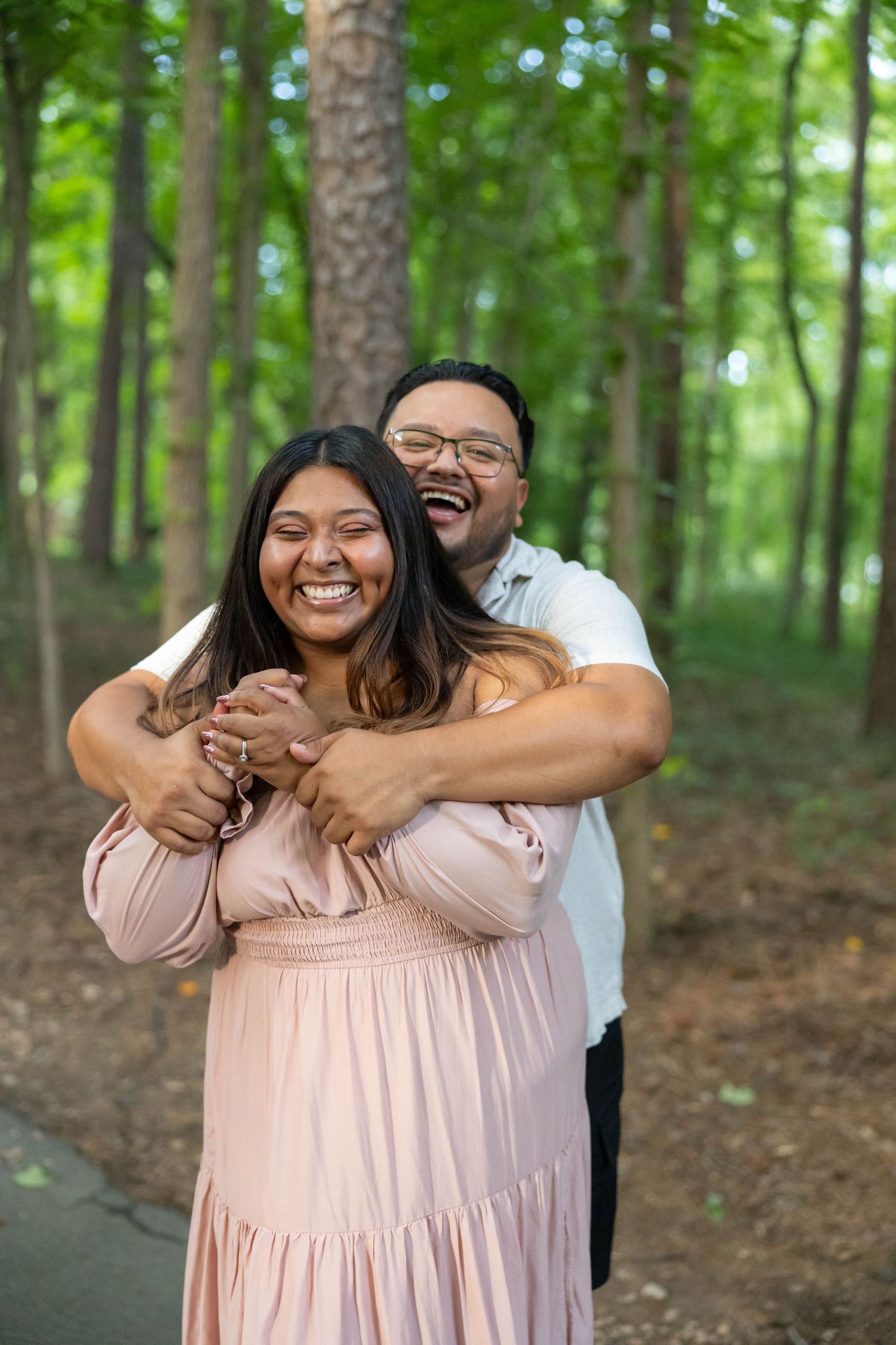 A joyful couple share a candid moment together while embracing in a lush forest setting, wearing pink dress and white shirt.