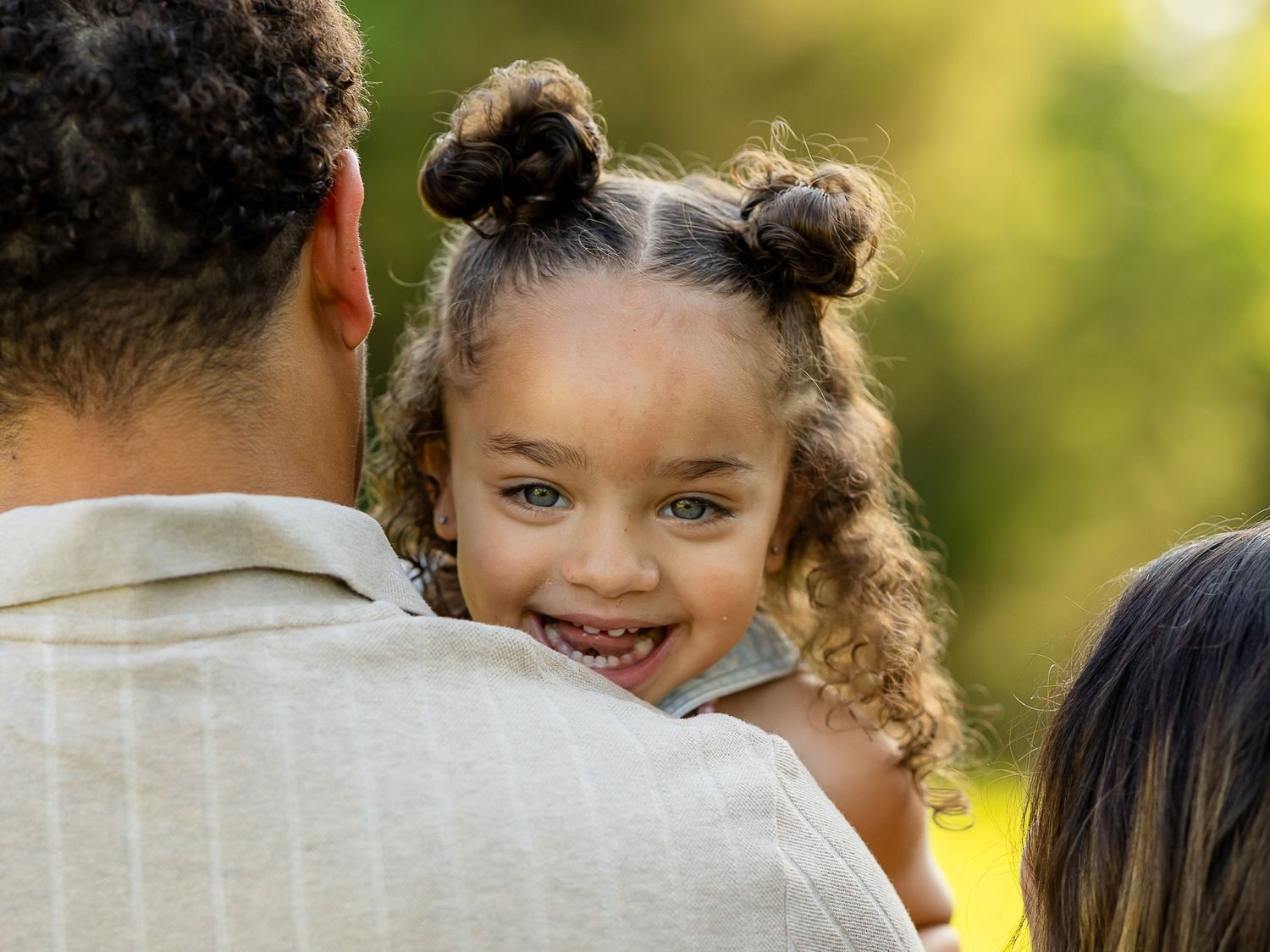 A smiling mixed-race toddler girl with bun pigtails being held by her father outdoors.
