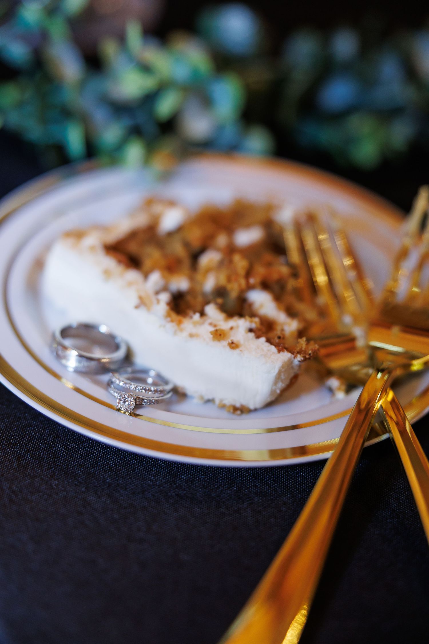 Wedding rings displayed beside a slice of cheesecake on a gold-rimmed plate with gold forks.