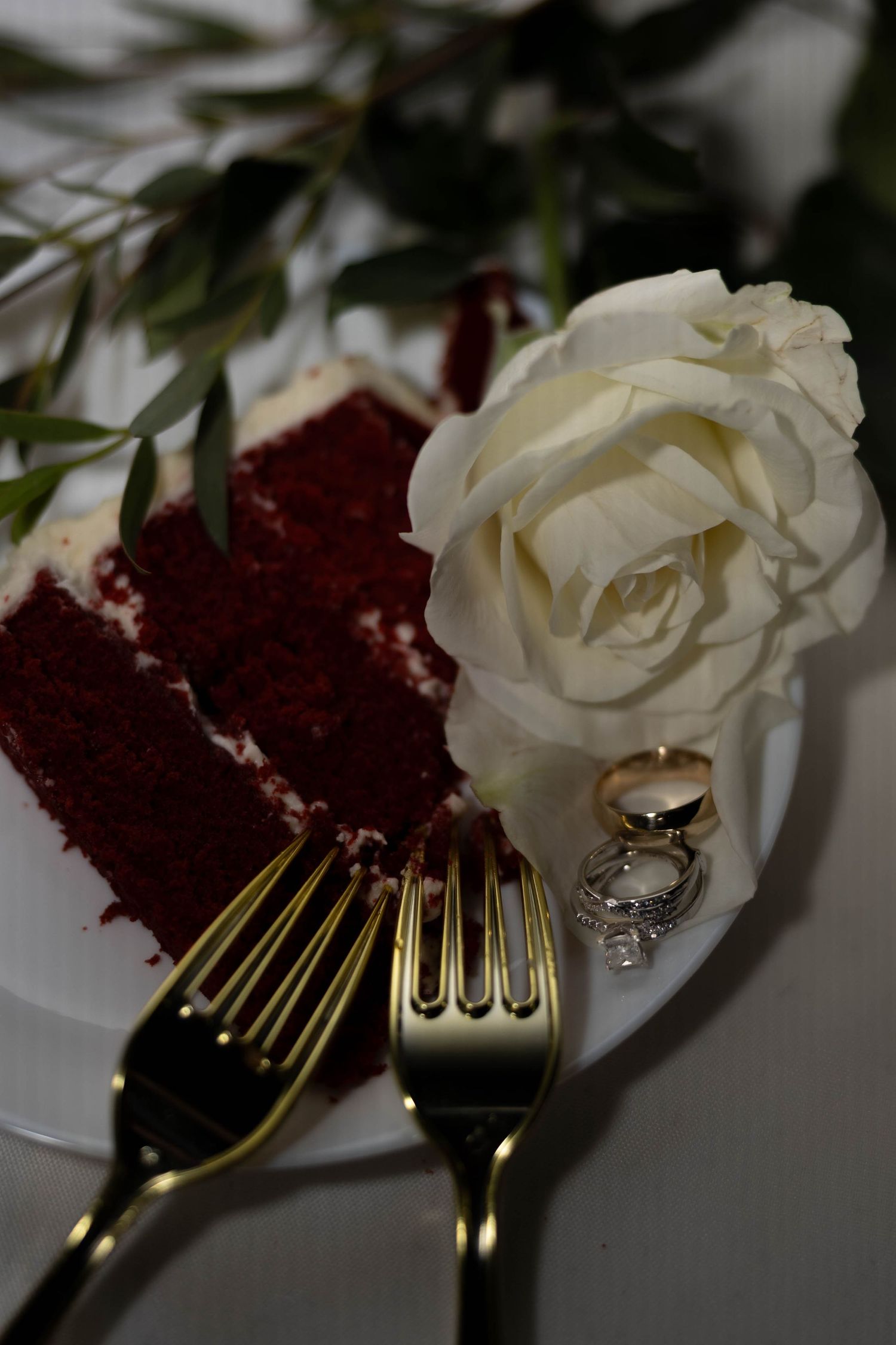 Red velvet wedding cake slice with white rose, gold forks, and diamond rings on white plate.