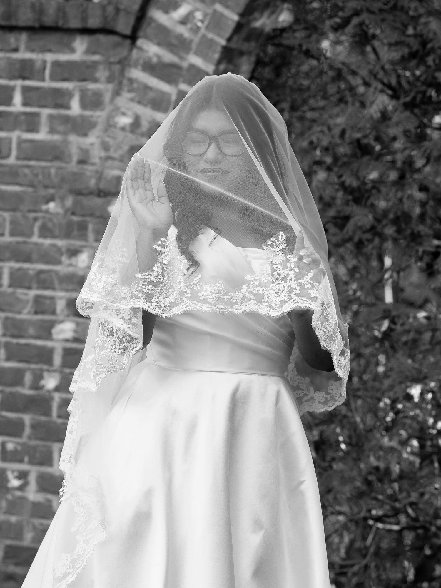 Bride in white wedding gown and lace veil with glasses poses outdoors in black and white photo.