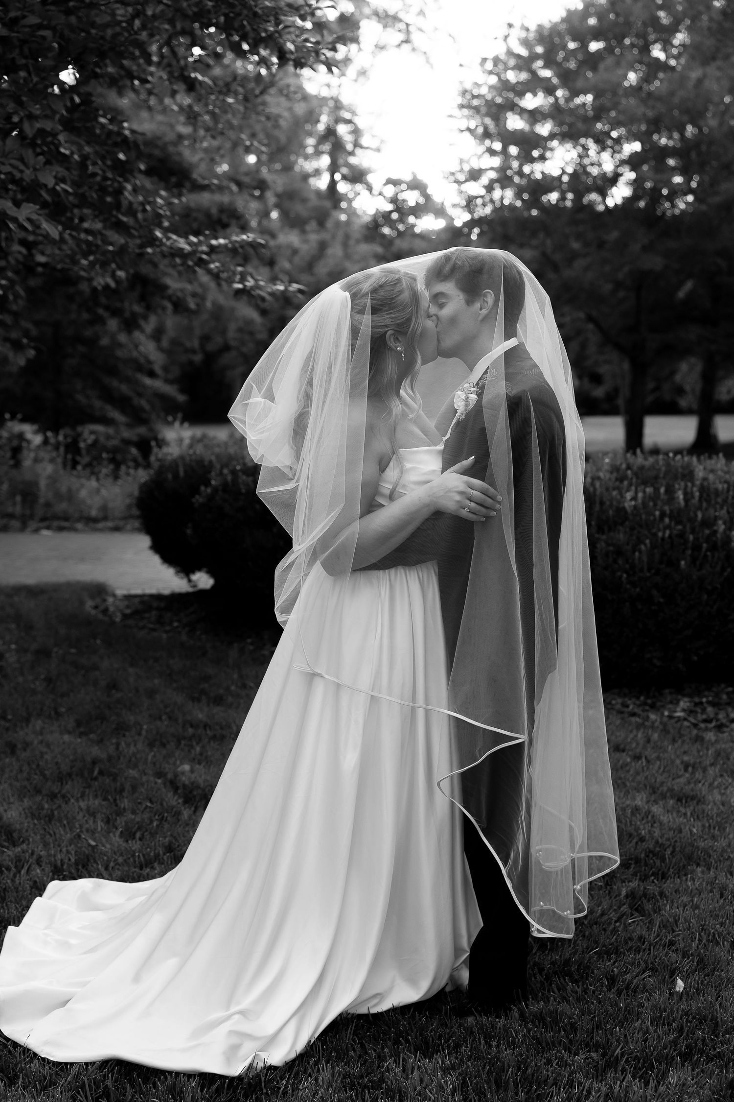 Black and white photo of bride and groom kissing under wedding veil in lush garden setting.