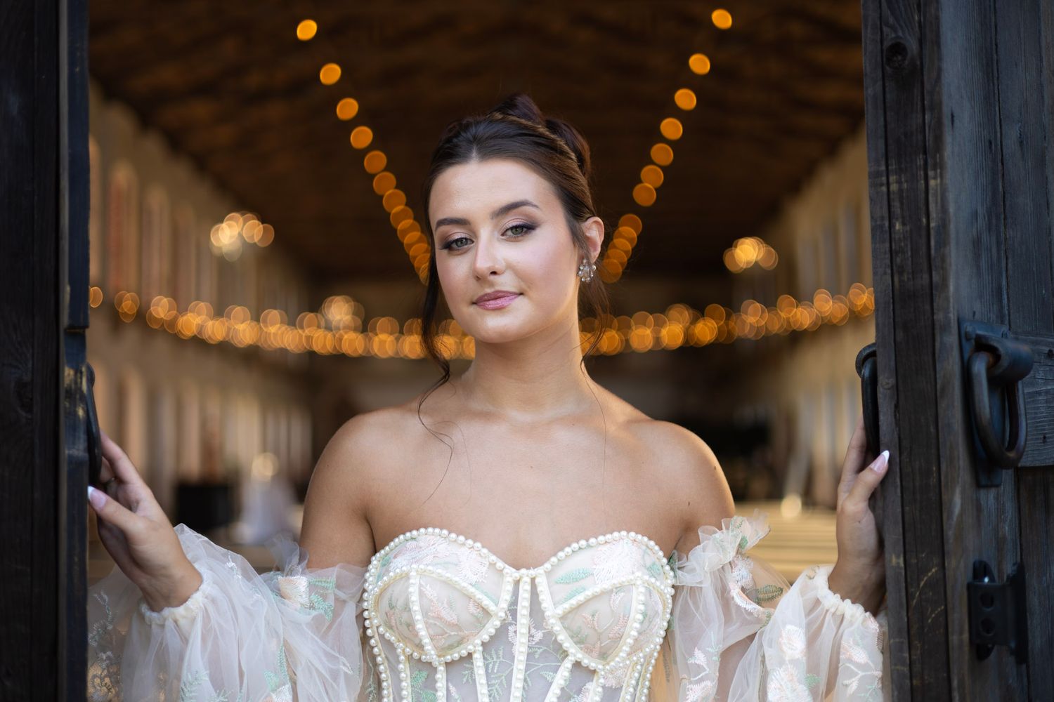 Elegant bride in white corset wedding dress and sheer sleeves poses in front of illuminated archway with string lights.