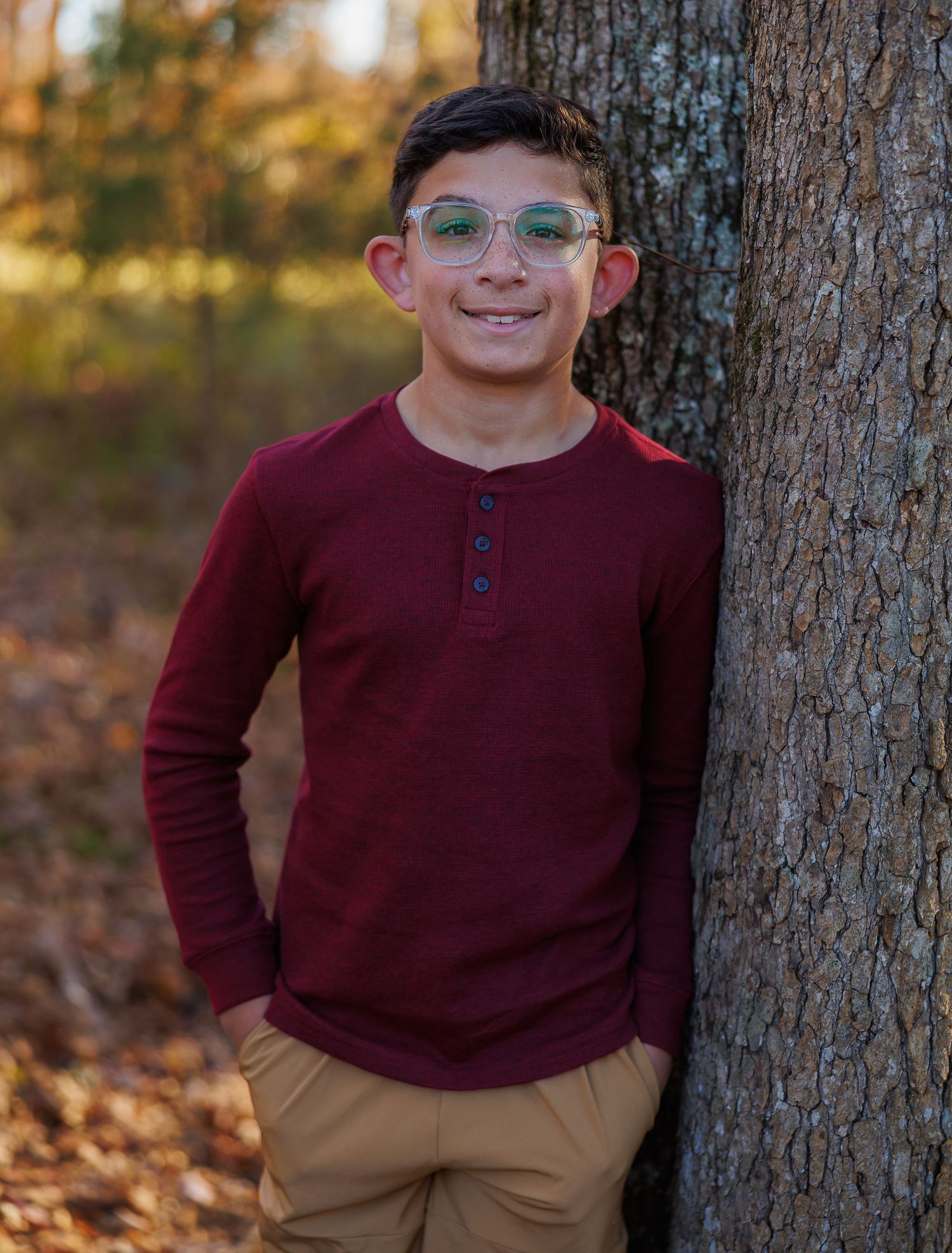 Child in a burgundy henley shirt and khaki pants poses casually against a tree trunk during autumn.