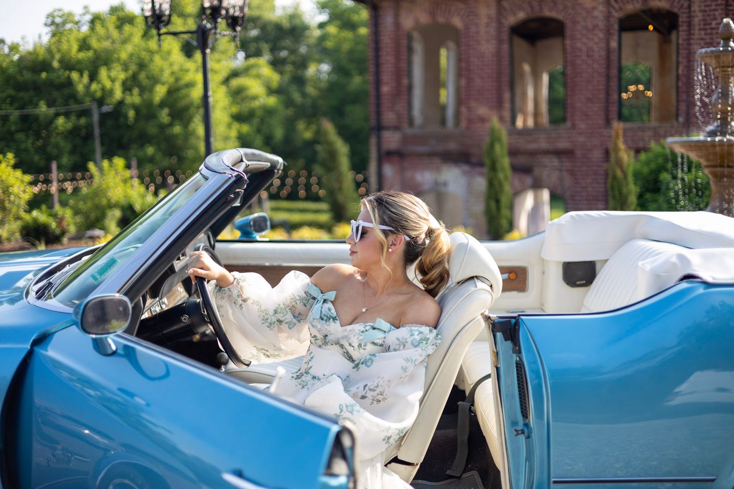 A classic light blue convertible car parked outdoors with people sitting inside during what appears to be a wedding.