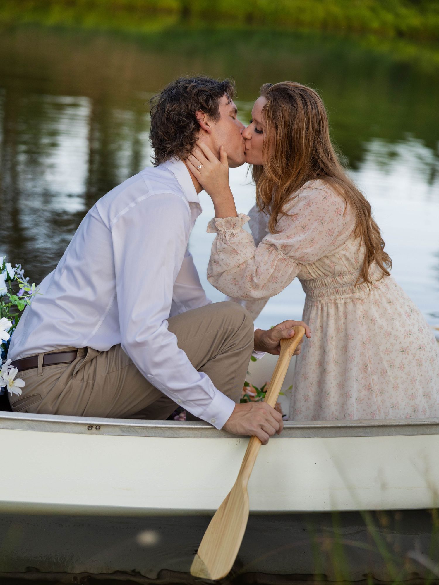 A romantic couple shares a tender kiss while sitting in a rowboat on a serene lake surrounded by greenery.