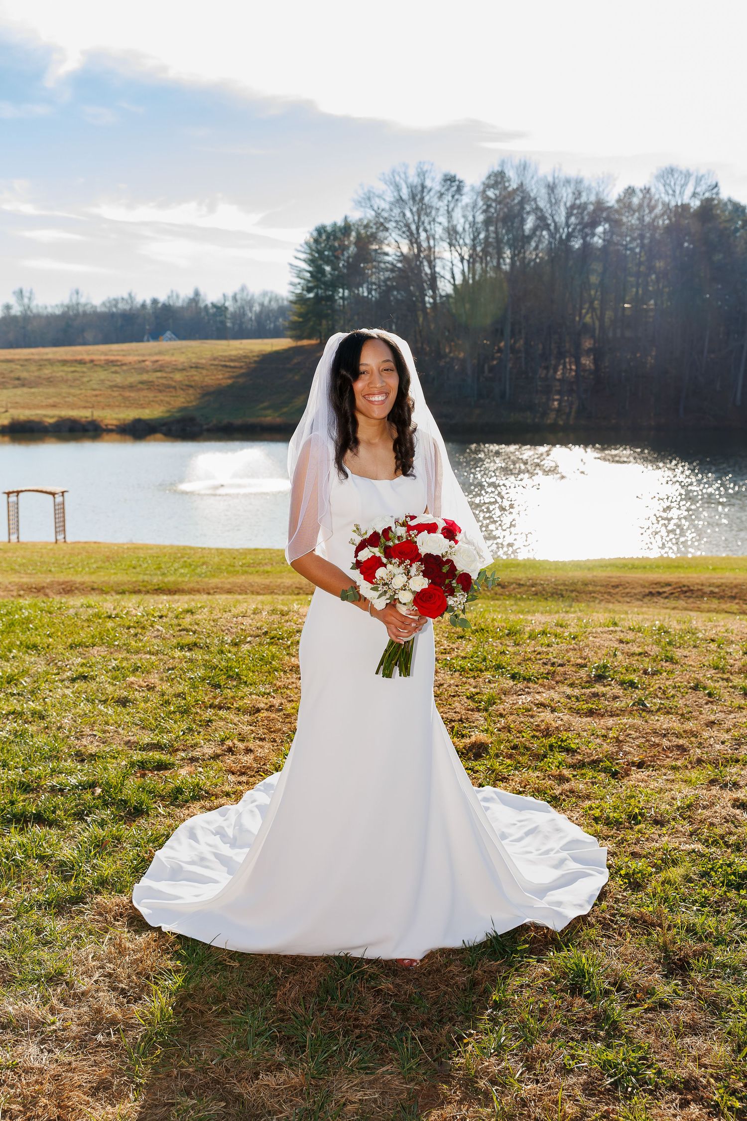 Smiling bride in white wedding gown and veil holding red and white bouquet by a serene pond.