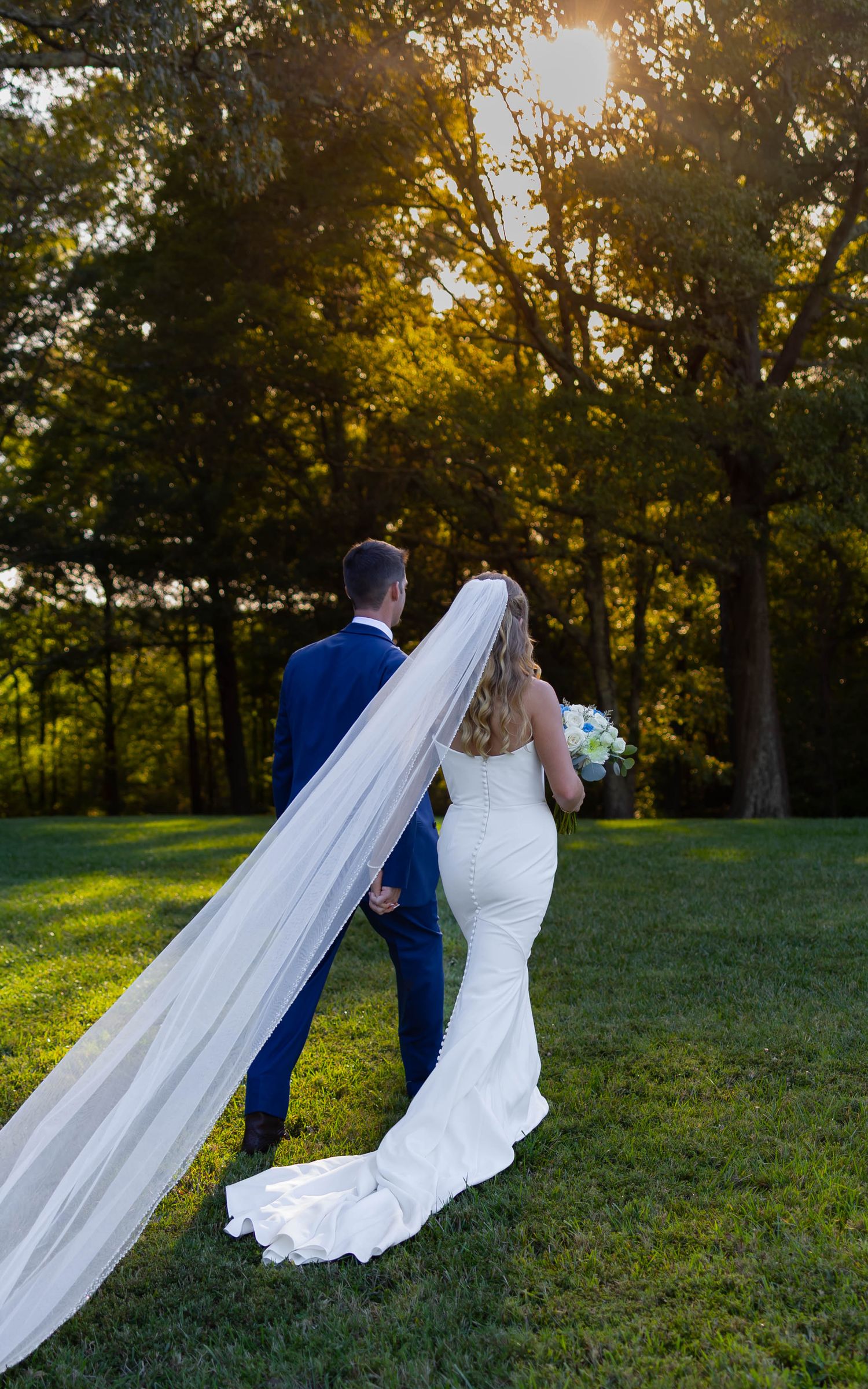 Bride in white gown with long veil and groom in blue suit holding hands on green lawn at golden hour.