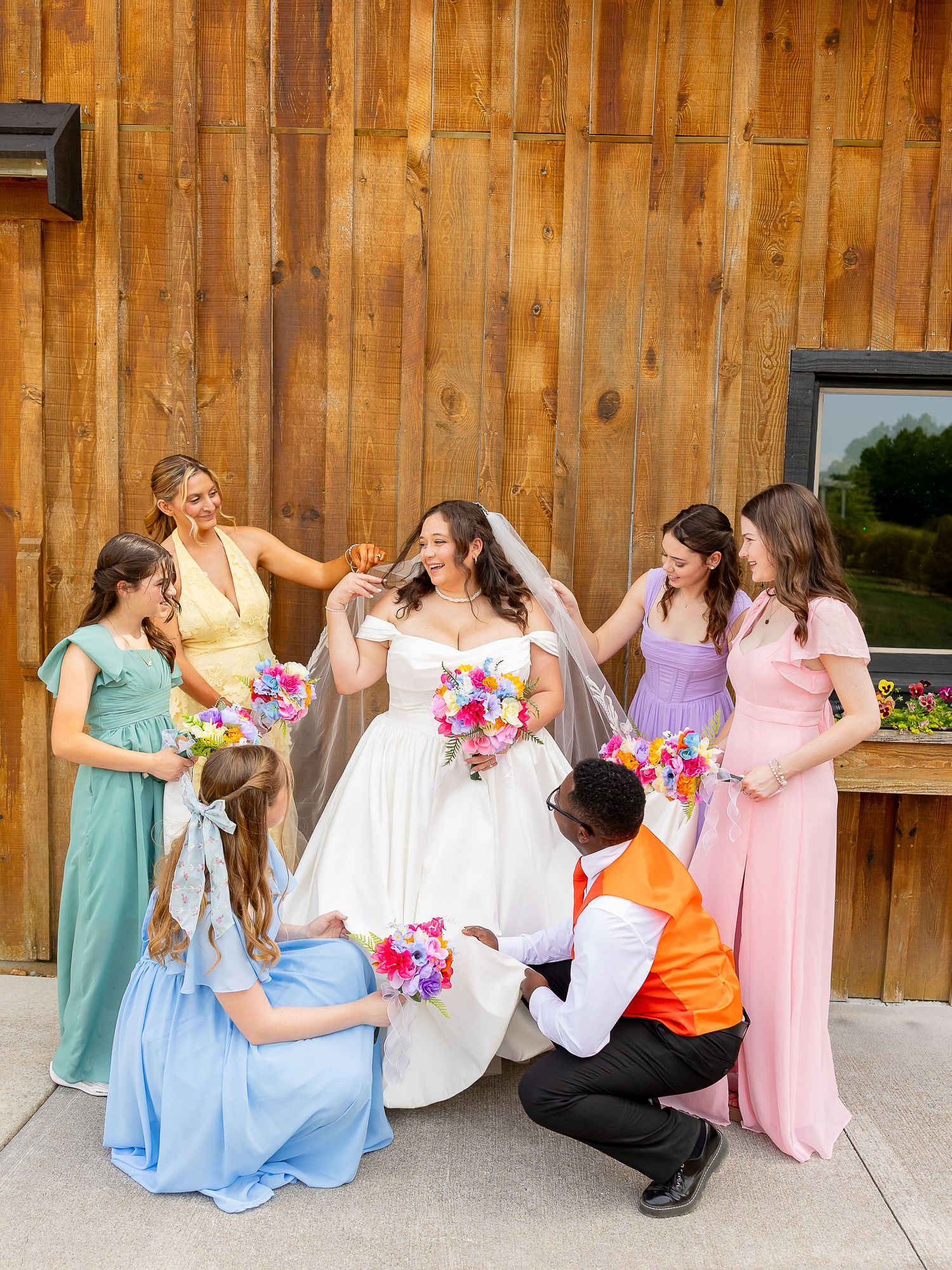 Bride in white gown with bridesmaids in colorful dresses adjusting her veil outside a rustic barn venue.