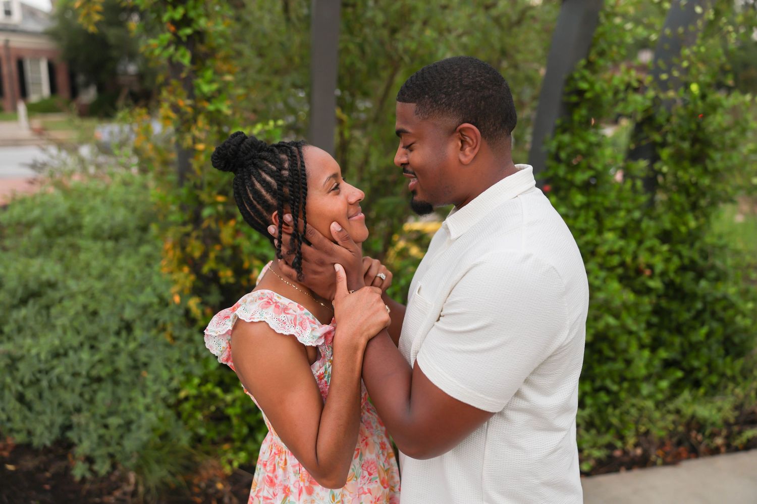 A couple shares a tender moment outdoors in front of greenery, with one wearing a floral dress and the other in white.