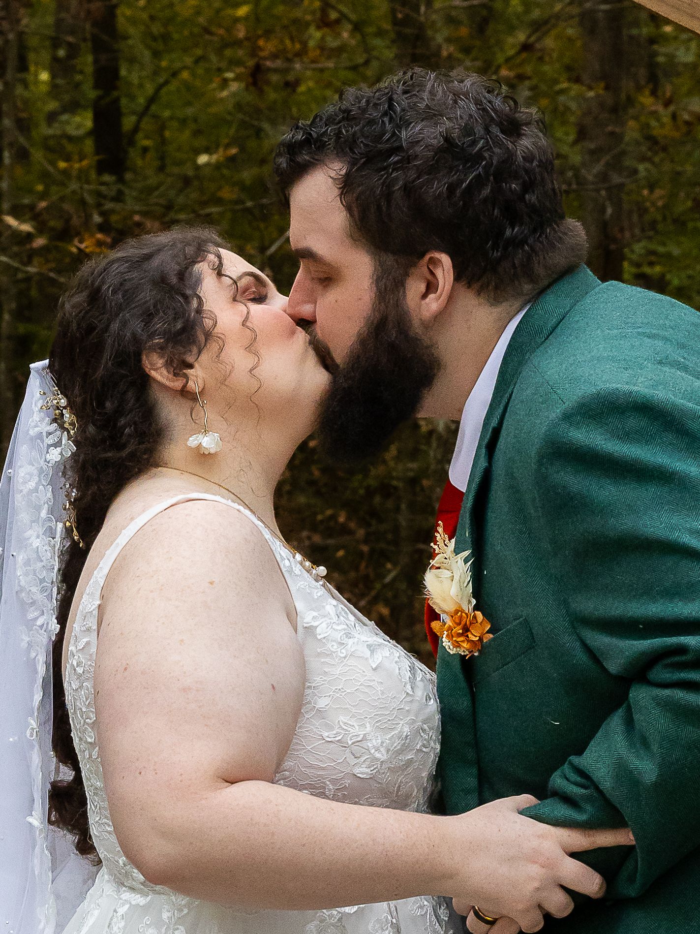 A bride in white lace dress and veil shares first kiss with bearded groom in green suit, fall foliage backdrop.