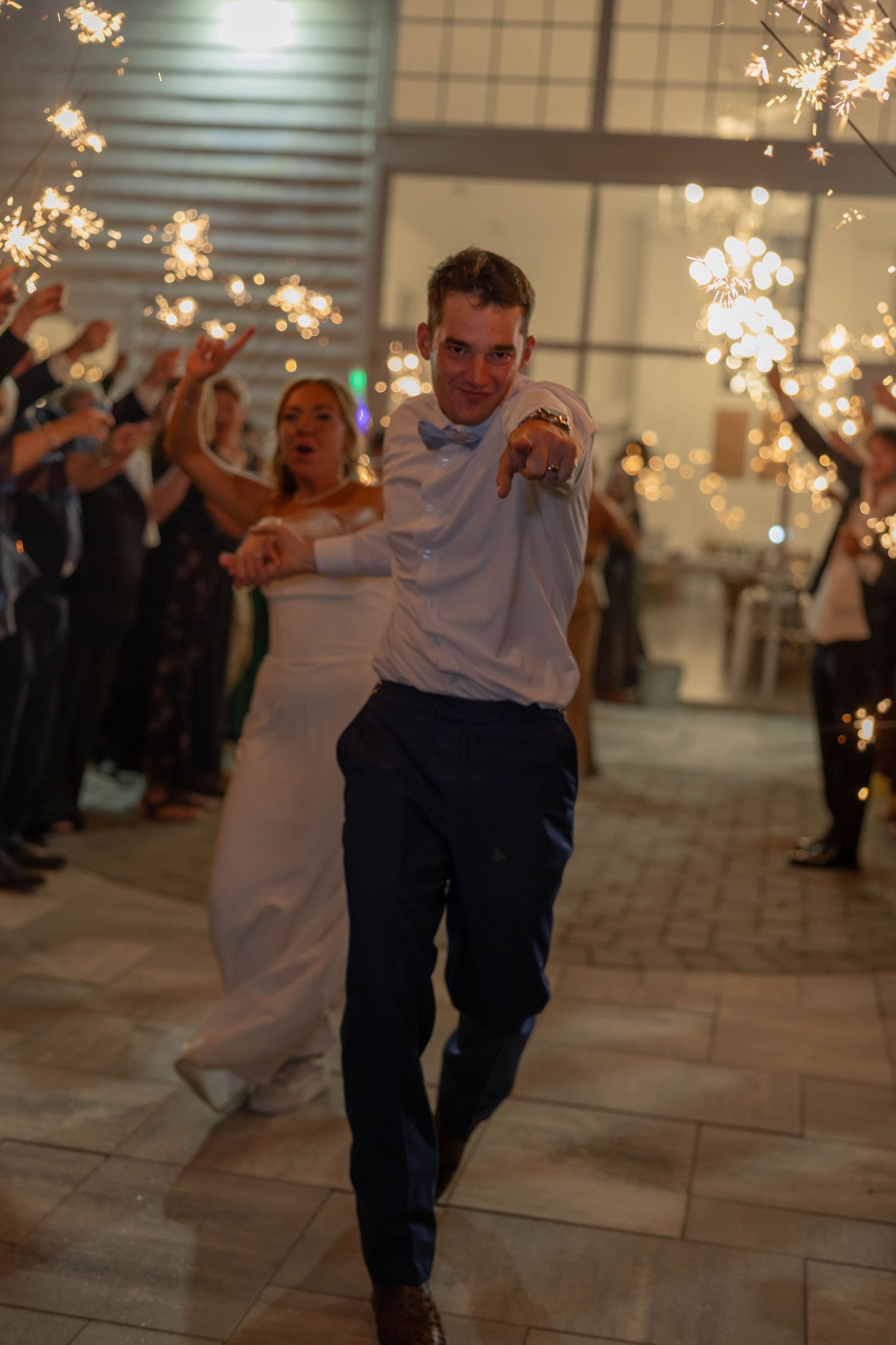 Bride and groom exit wedding reception through sparkler tunnel, groom pointing at camera, guests lining the aisle.