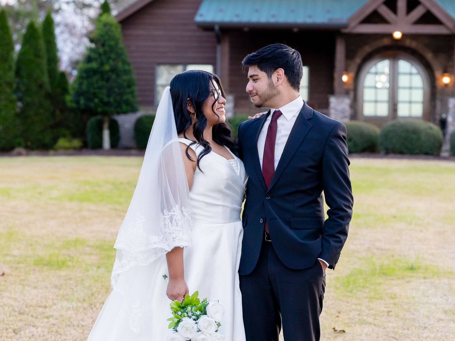 Bride and groom in navy suit smiling at each other on wedding day outside rustic chapel.