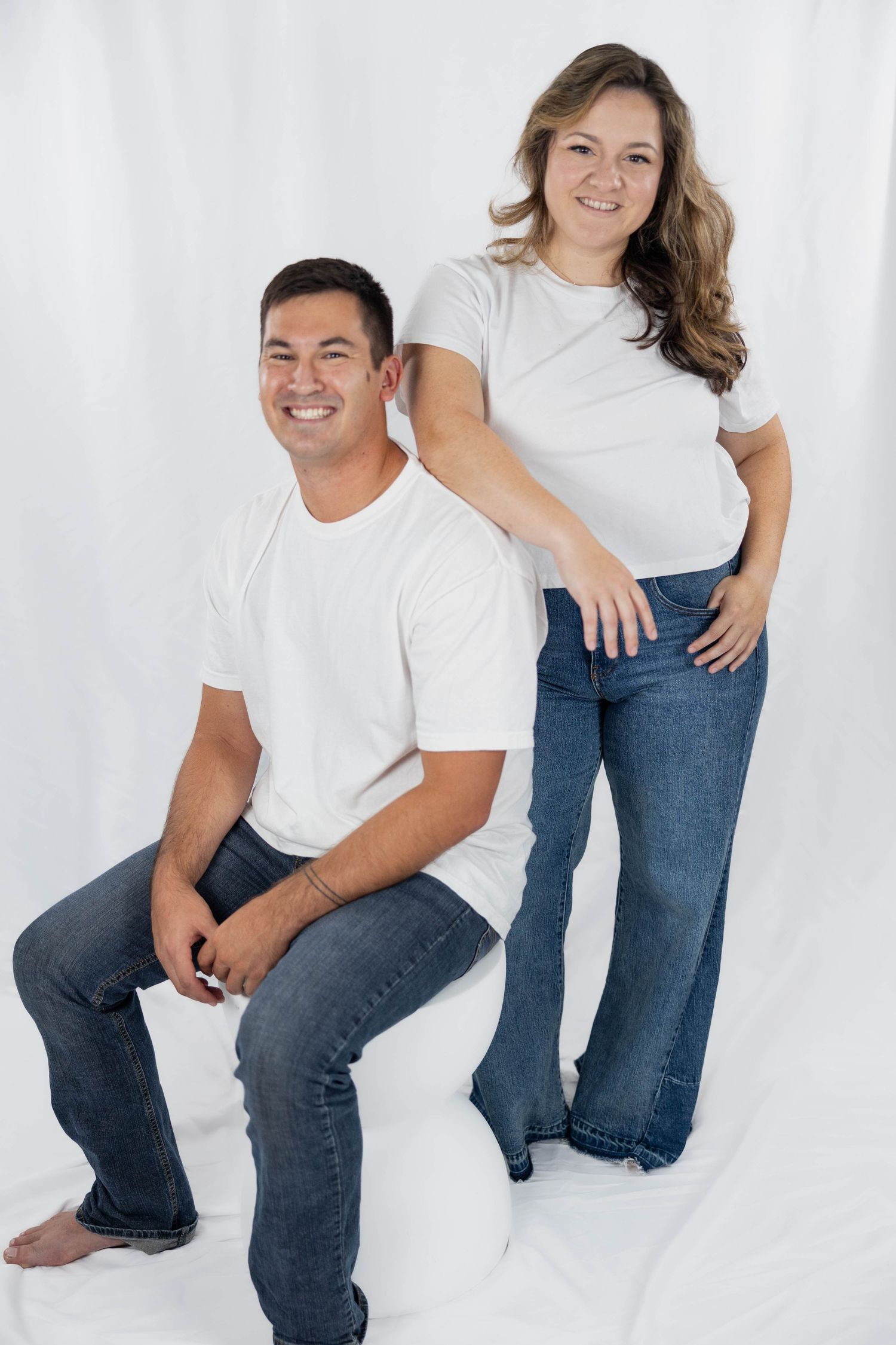 A couple wearing matching white t-shirts and blue jeans pose together against a white backdrop in a casual studio setting.