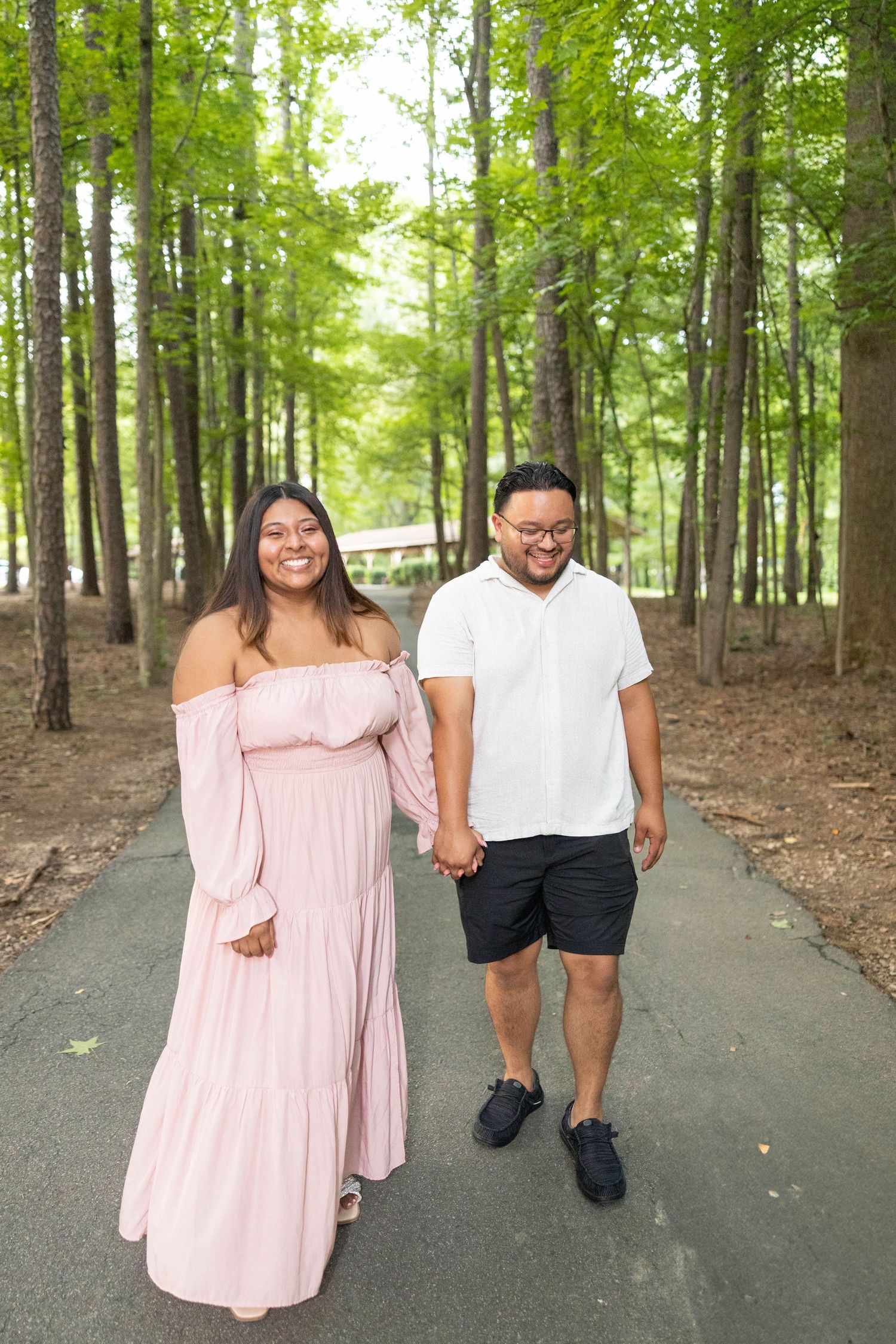 A couple walking hand in hand on a paved path through a serene wooded forest on a bright summer day.