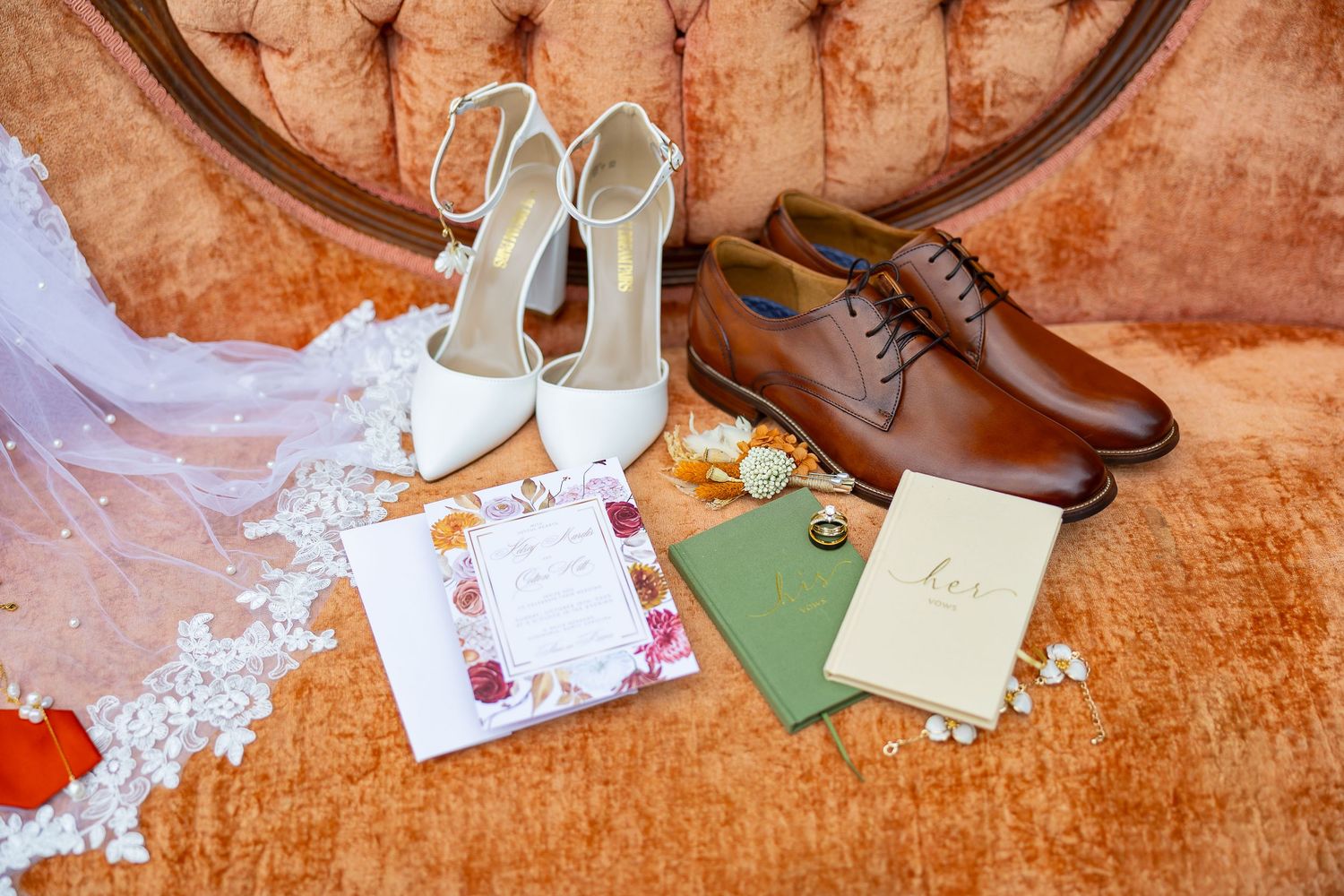 White bridal heels and brown groom dress shoes displayed with wedding invitations and green envelopes on a velvet chair.