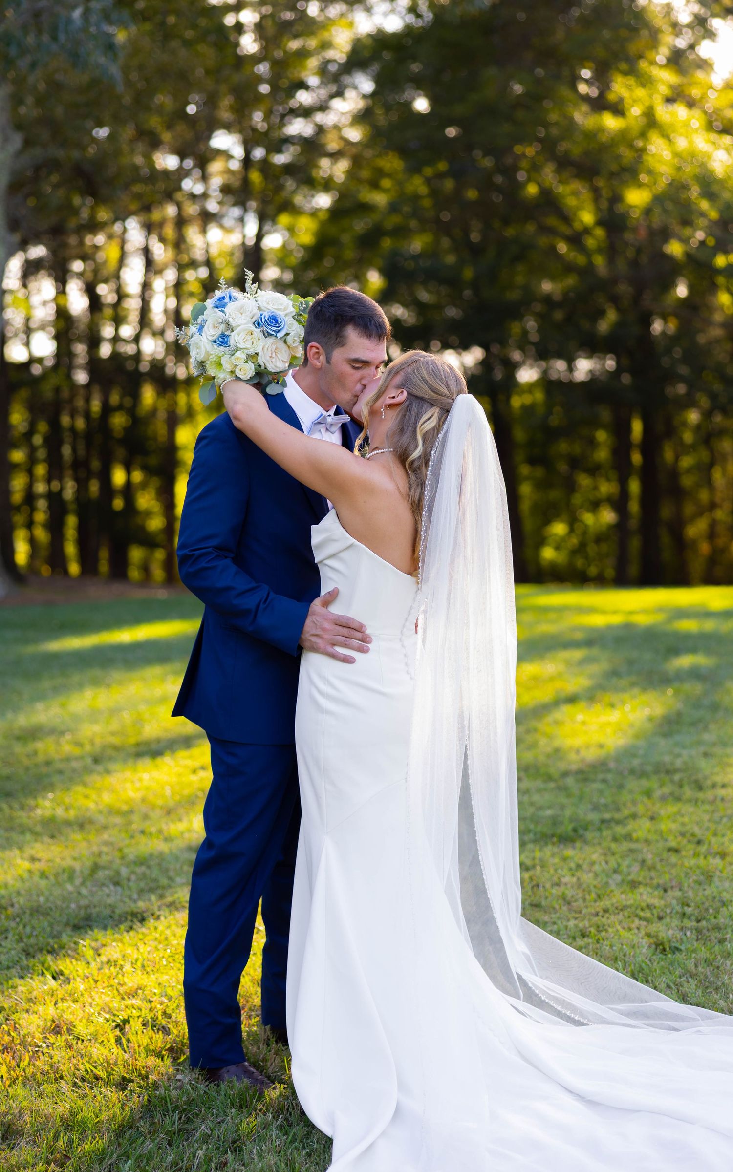 Bride and groom sharing a kiss on green lawn, she in white gown with veil, he in navy suit, holding bouquet.