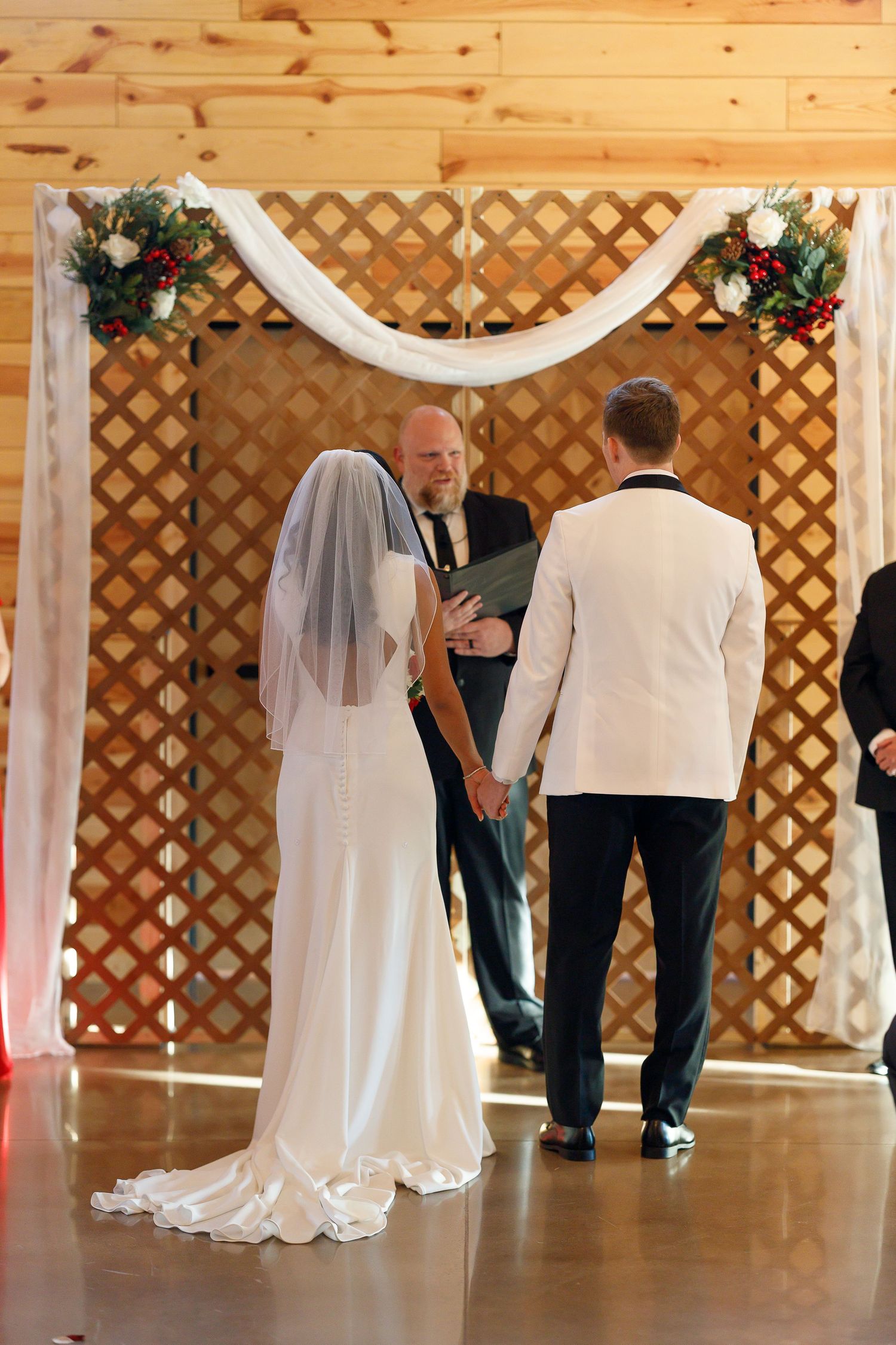 Bride and groom exchange vows before officiant at rustic wooden lattice altar in barn wedding venue.