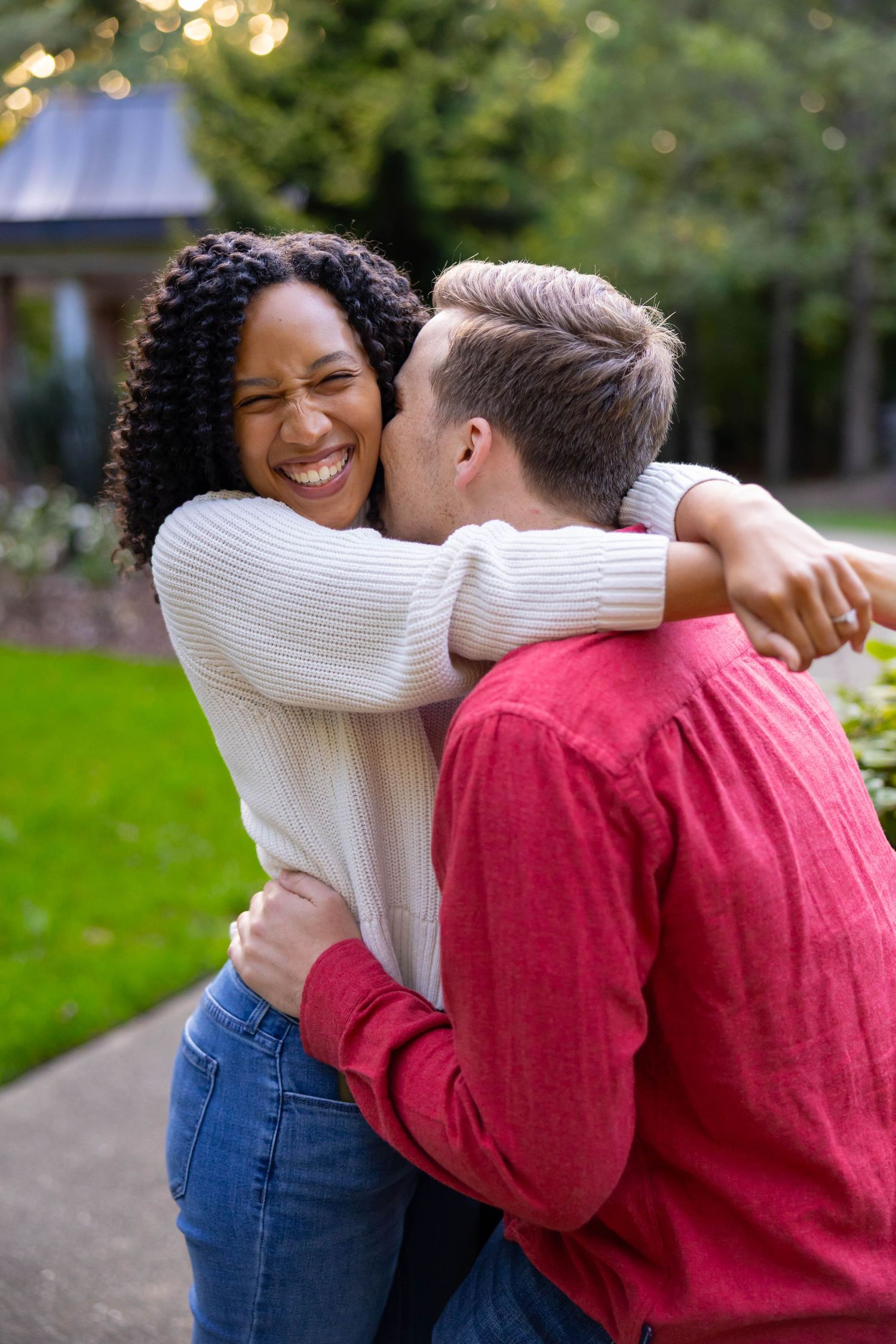 A couple embraces outdoors on a sunny day, wearing a white sweater and red shirt against a natural background.